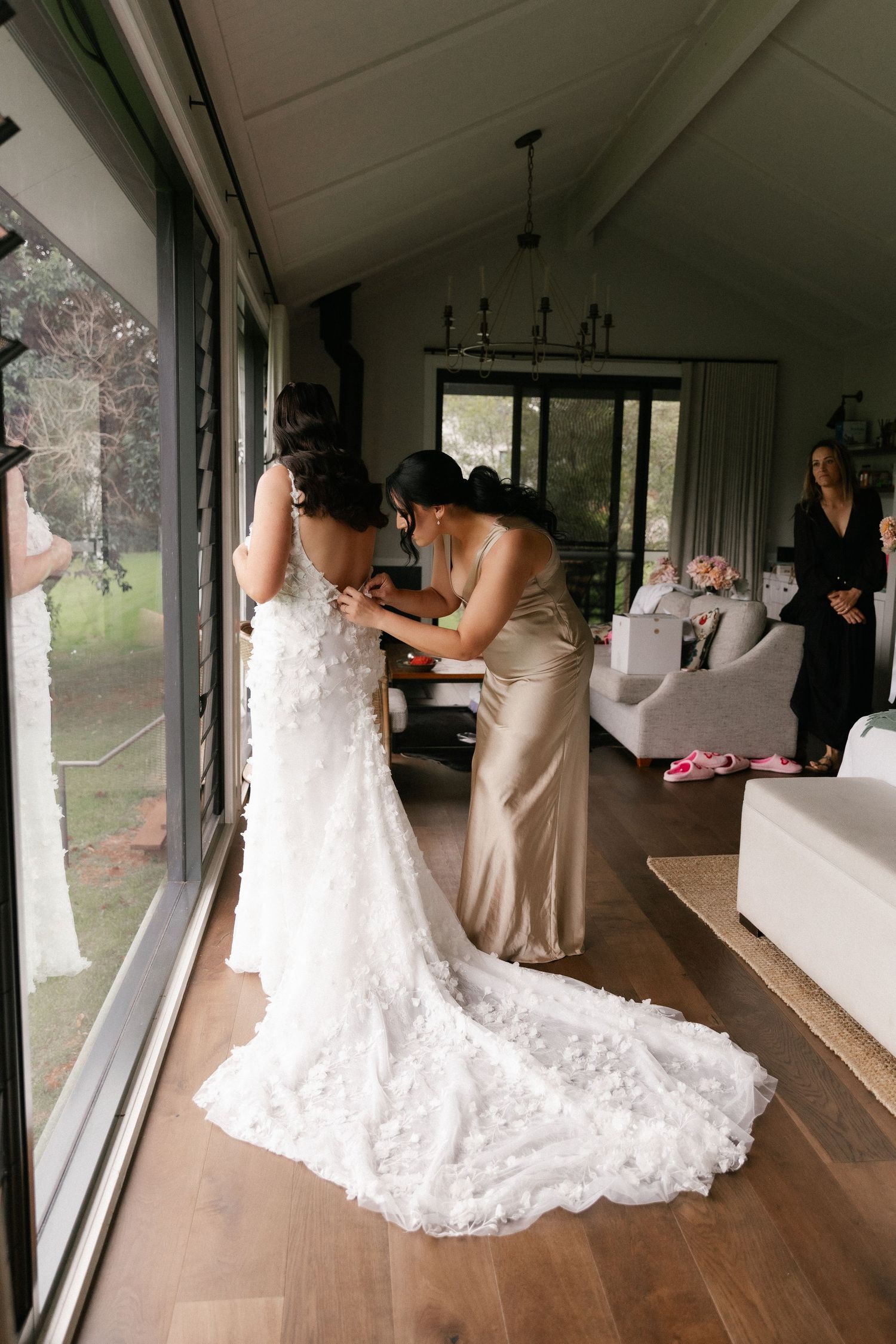 A bridesmaid in a gold dress helps a bride in a floral lace gown with her wedding dress in an elegant venue.