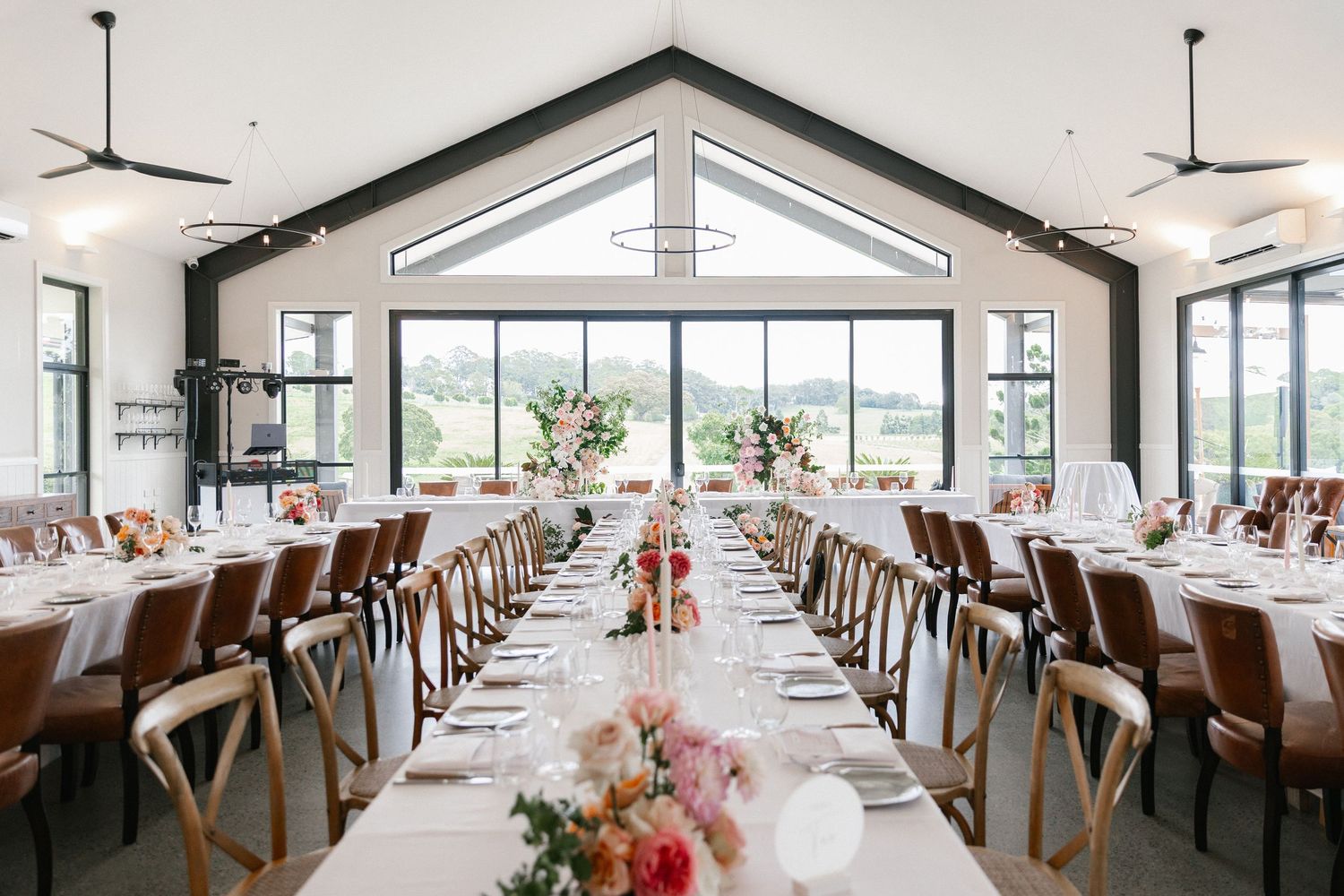 Elegant wedding reception hall with long tables, leather chairs, and large windows overlooking greenery.