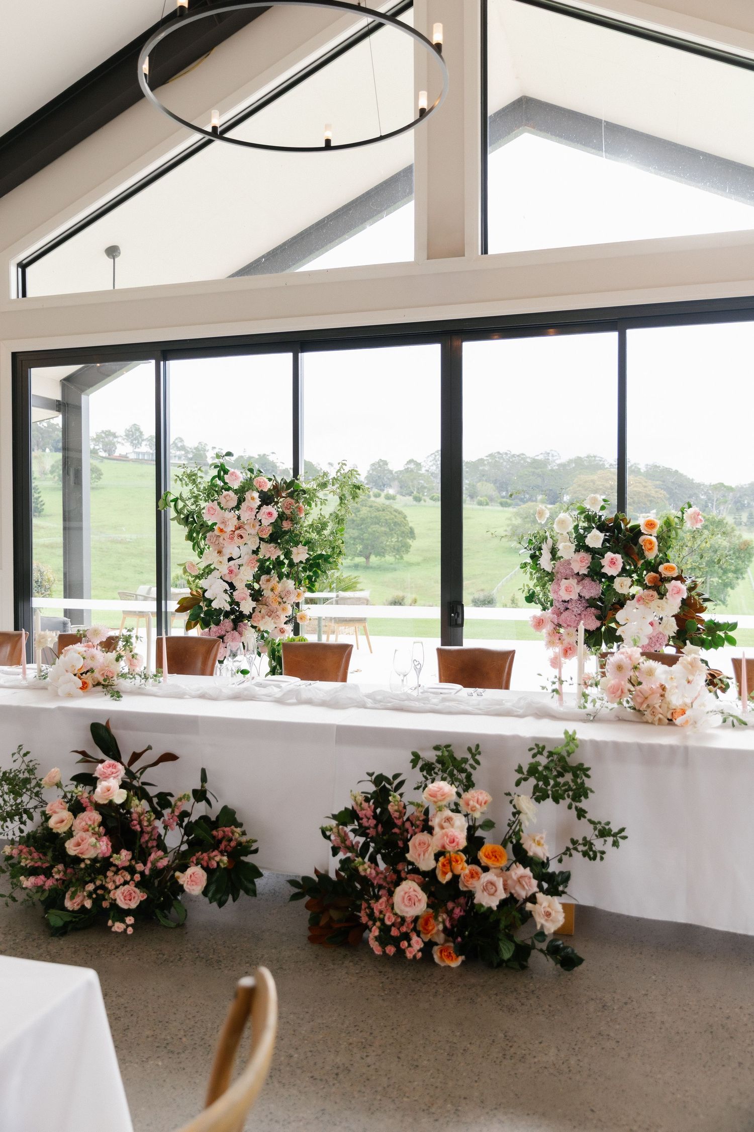 Elegant wedding reception venue with long white table adorned with tall and low pink floral arrangements by large windows.