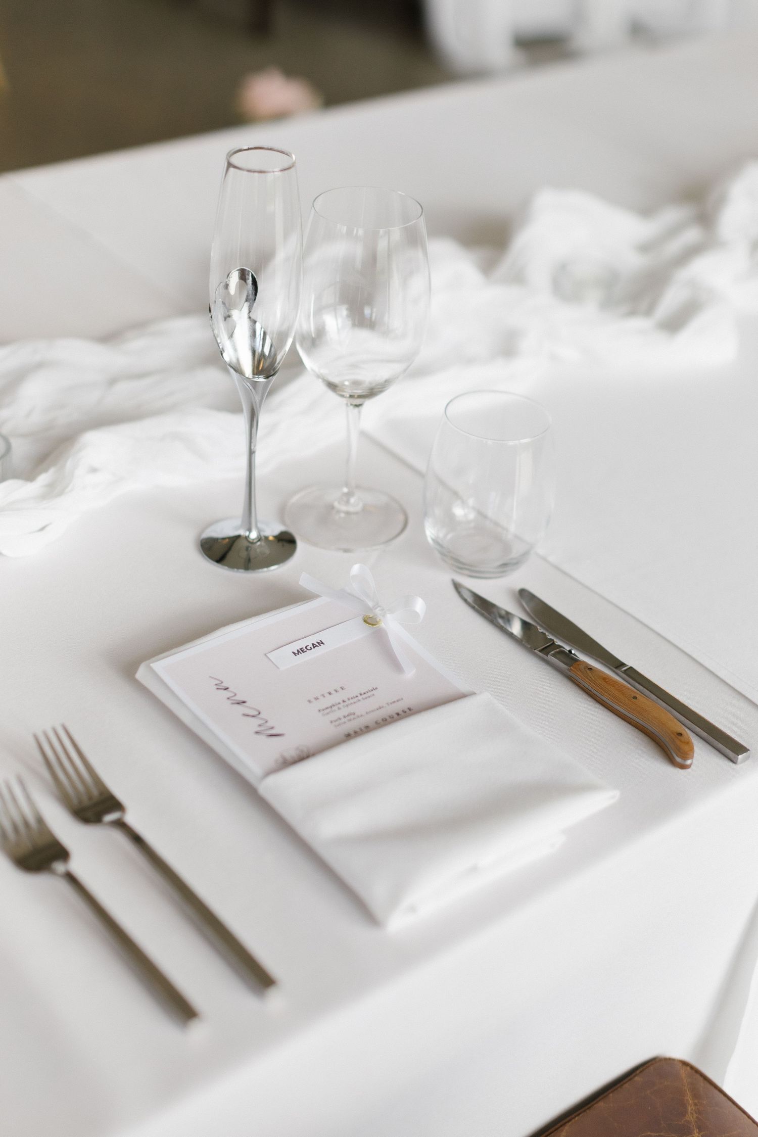 Elegant white wedding table setting with crystal glassware, silverware, and minimalist menu card.