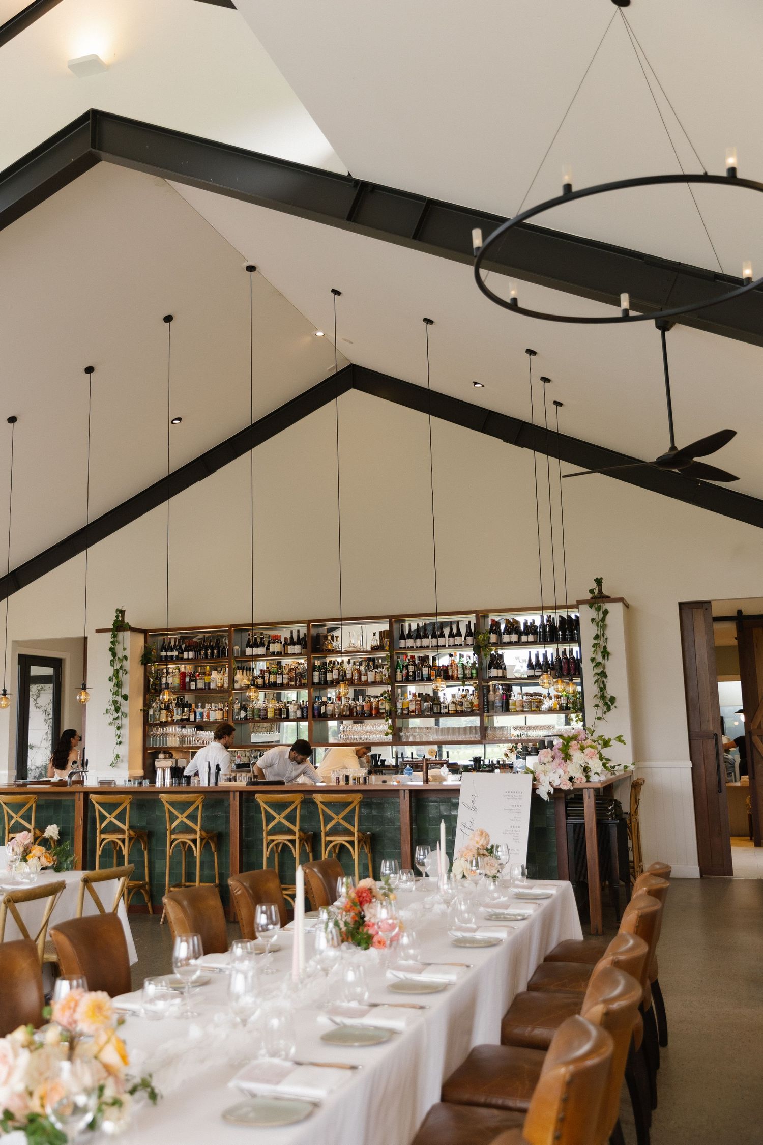 Elegant wedding reception venue with high vaulted ceilings, long white tables, leather chairs, and a fully stocked bar backdrop.