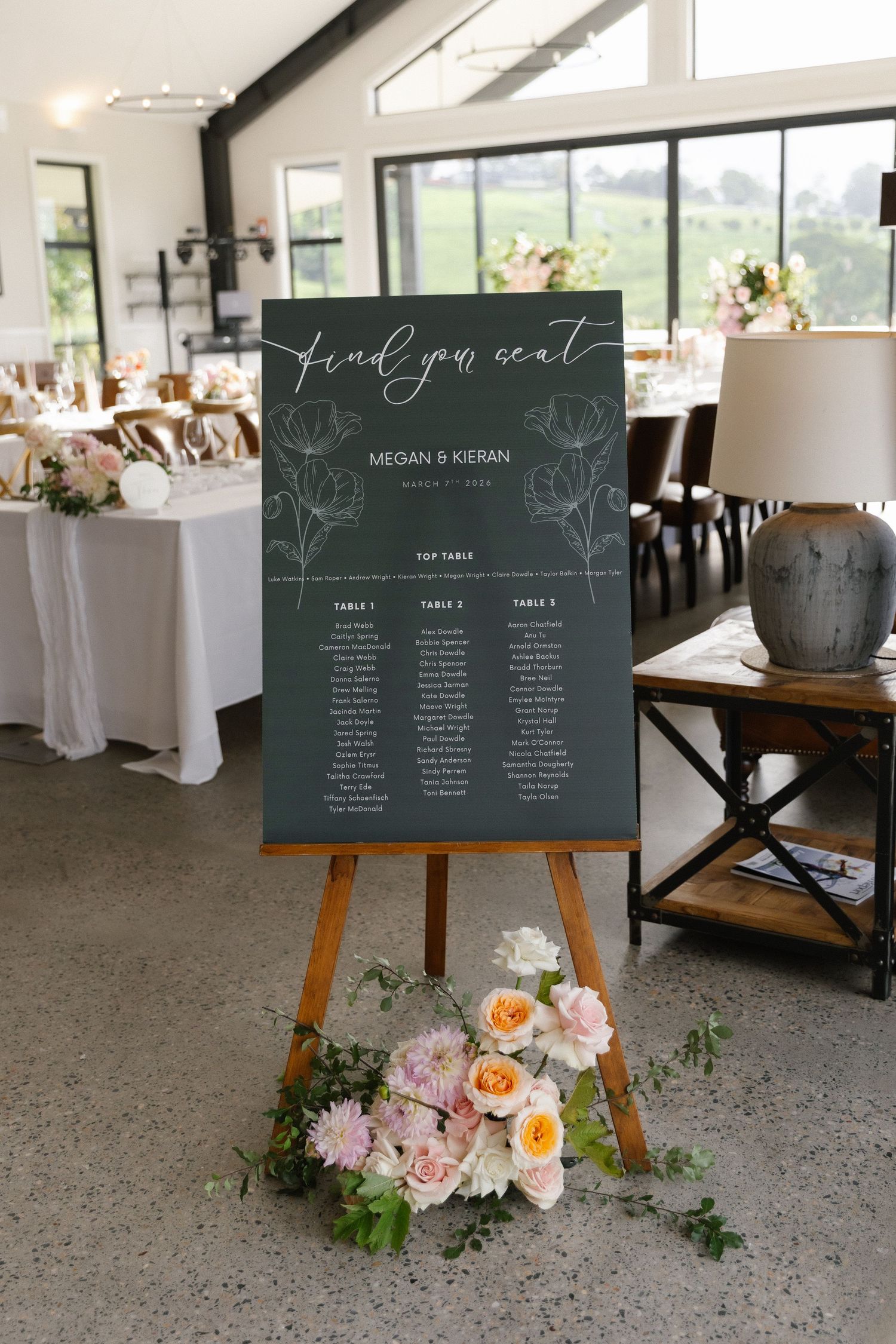Dark green wedding seating chart on an easel with floral arrangement below, reading Find Your Seat for Megan and Kieran.