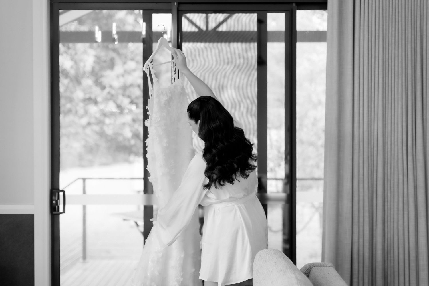 Black and white photo of a bride in a robe holding her wedding dress near a window.