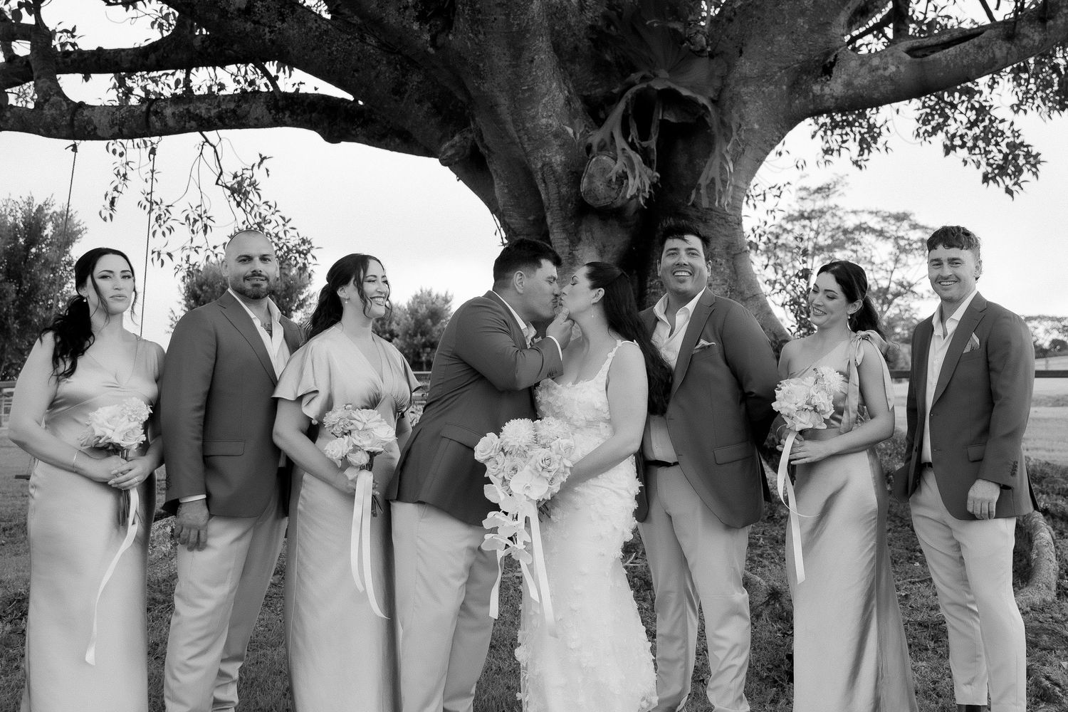 Black and white wedding party photo of bride and groom kissing surrounded by bridesmaids and groomsmen under large tree.