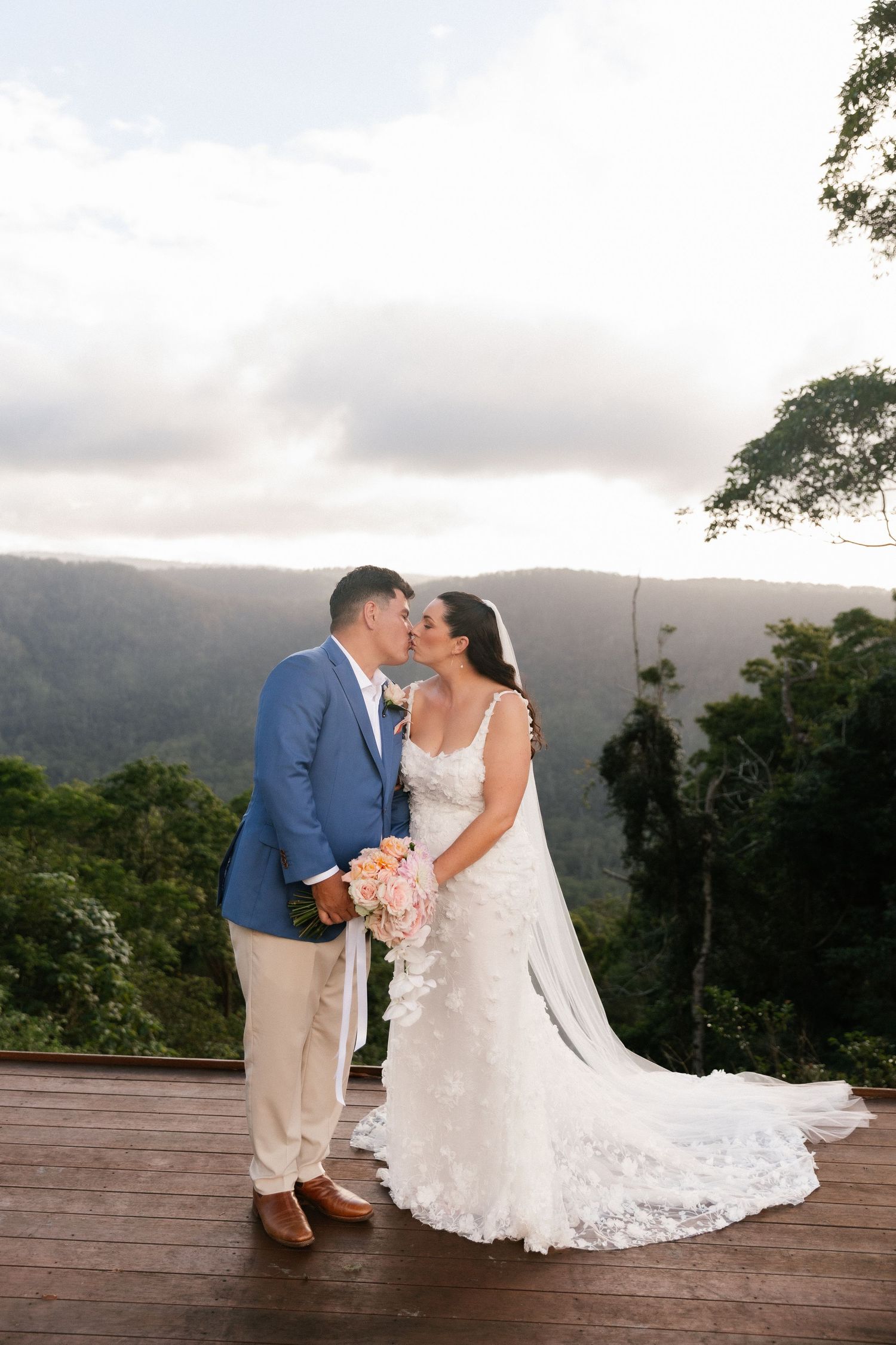 Bride and groom share a kiss on a wooden deck overlooking lush mountain forest at sunset wedding.