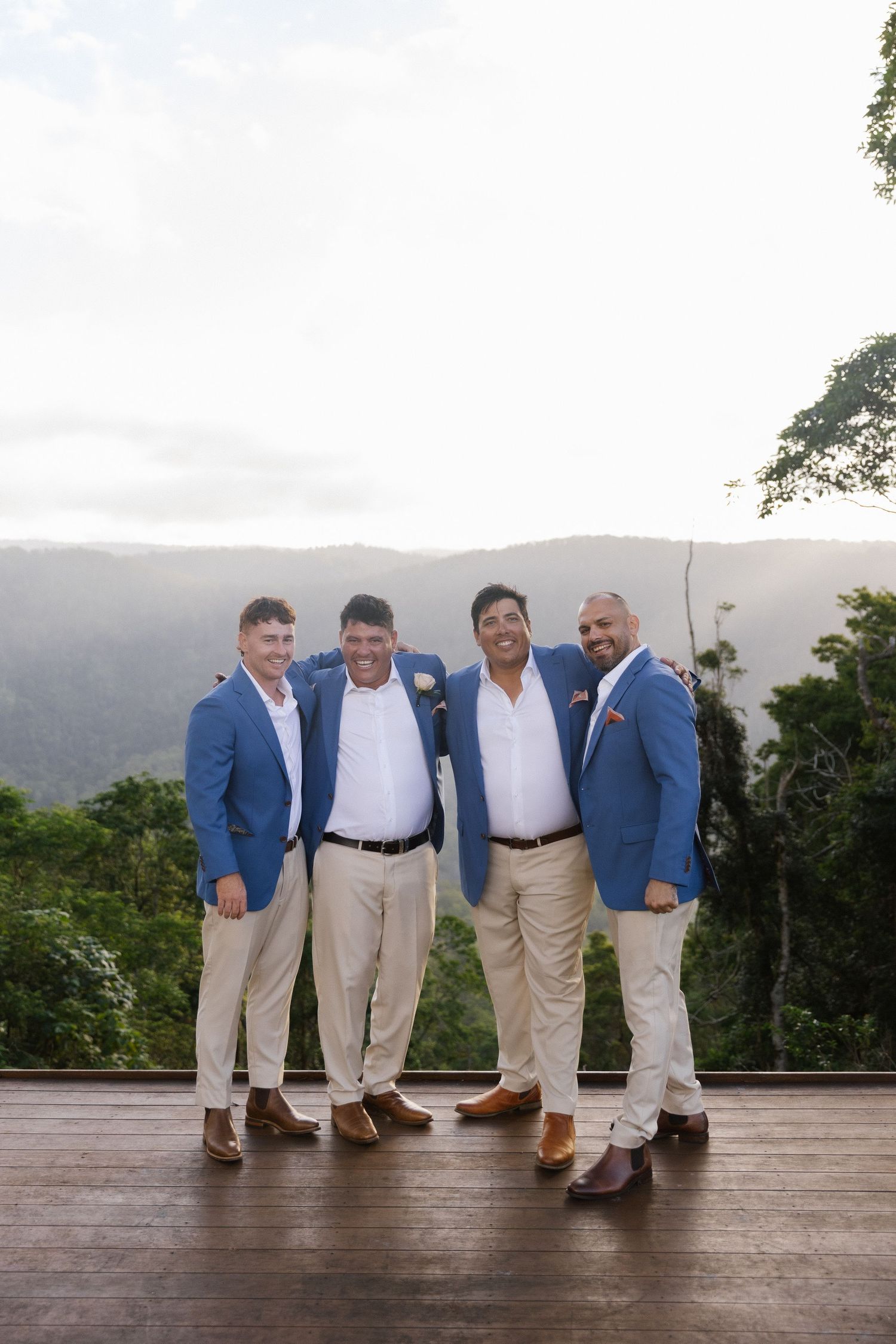 Groom and three groomsmen in matching blue blazers posing on a deck with mountain views behind them.