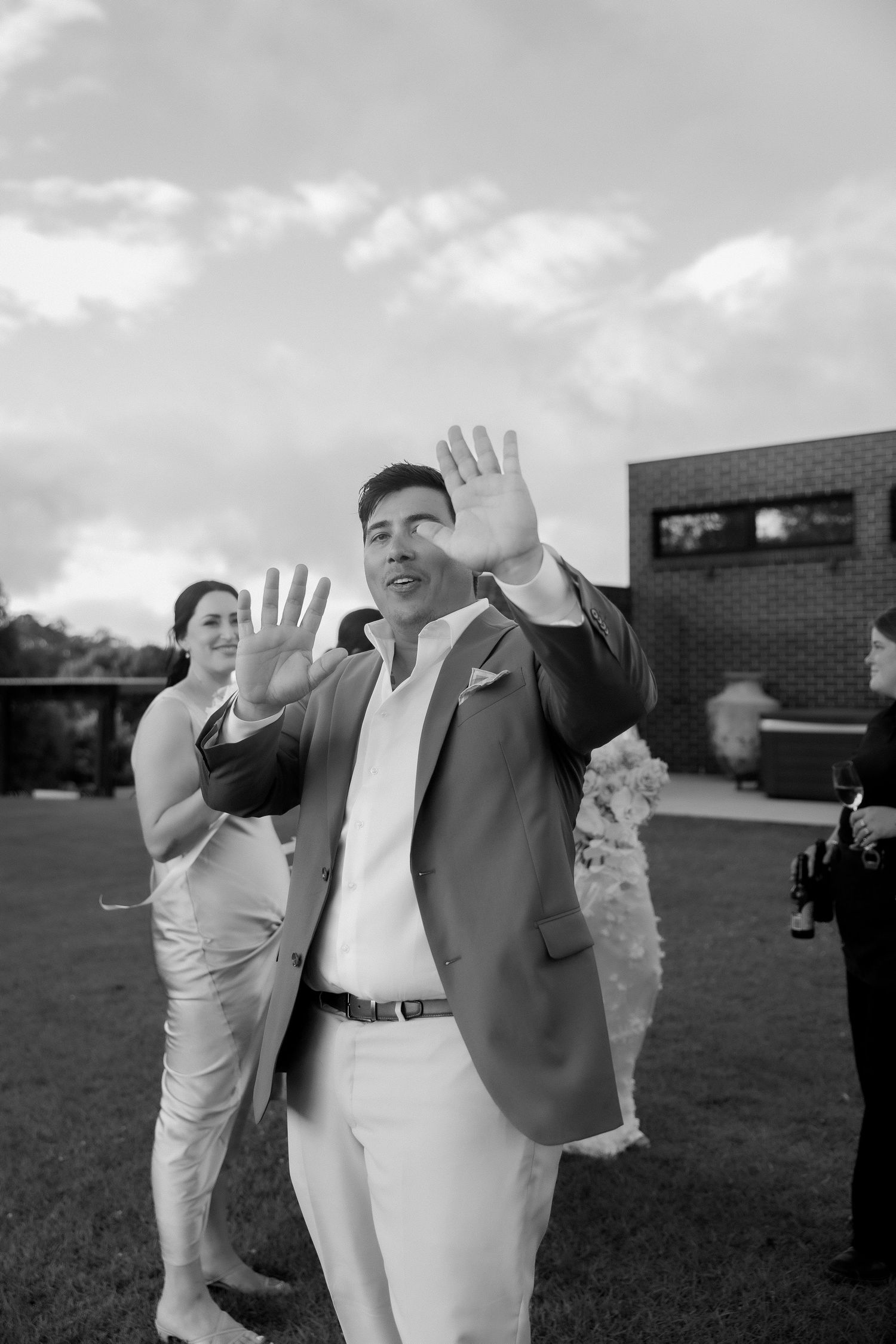 Black and white photo of groom in suit waving both hands at outdoor wedding celebration.