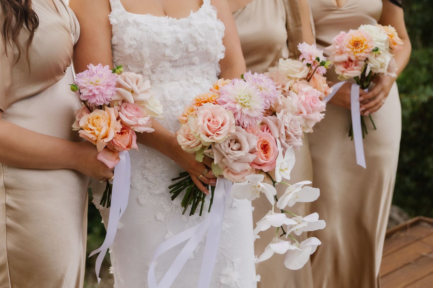 Bride in white lace gown and bridesmaids in champagne dresses holding pink and peach wedding bouquets with ribbons.