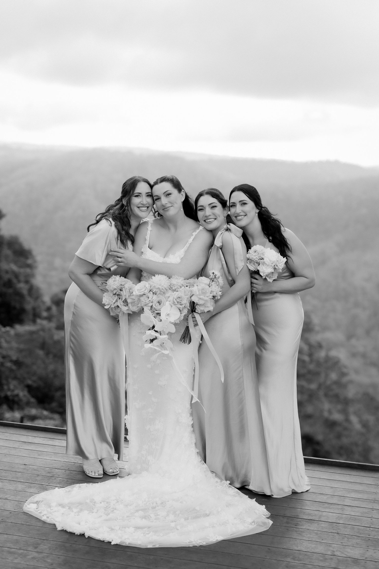 Bride and three bridesmaids in elegant gowns pose together on a deck with mountain views in black and white.