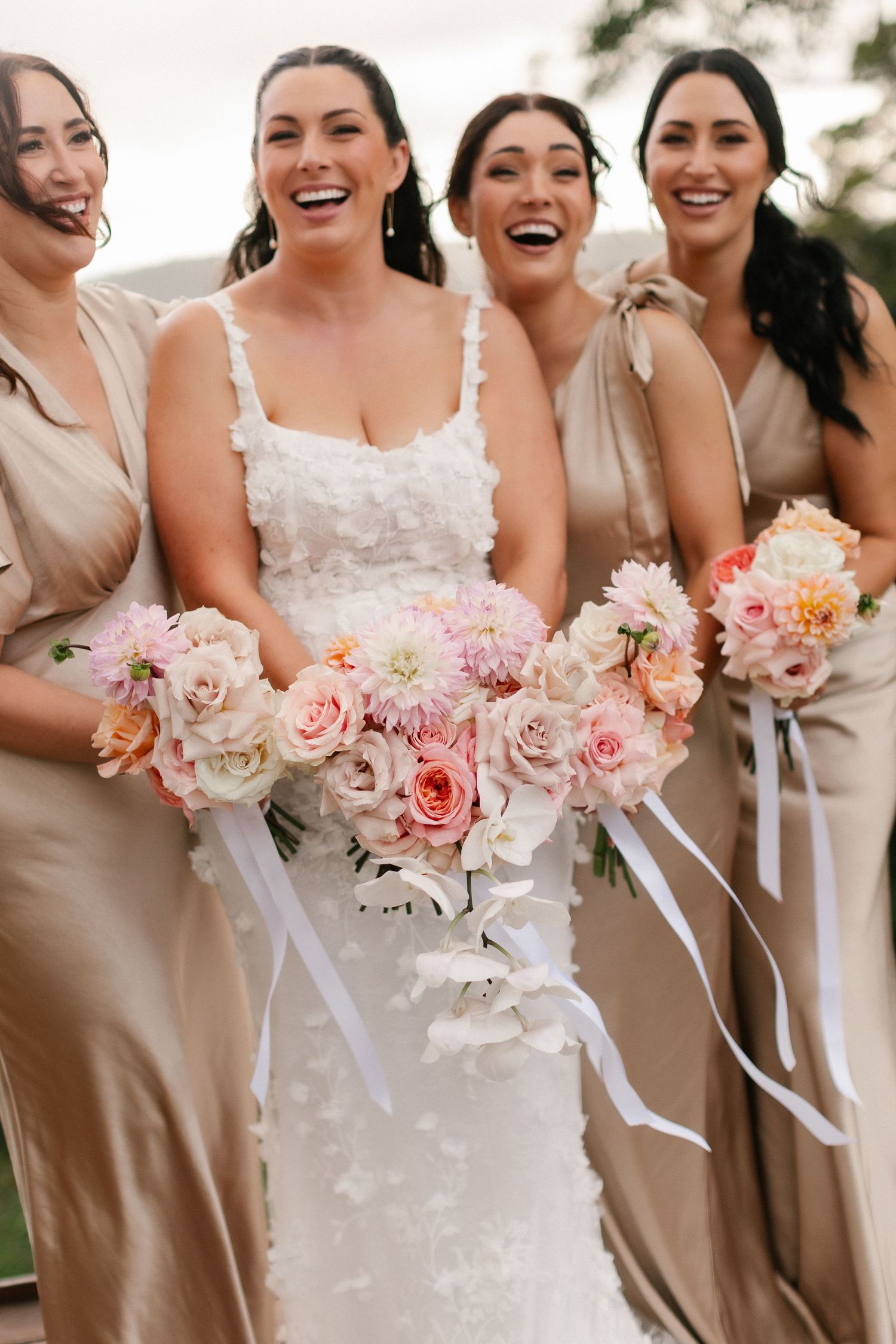 Bride in white floral gown laughing with four bridesmaids holding pink bouquets.