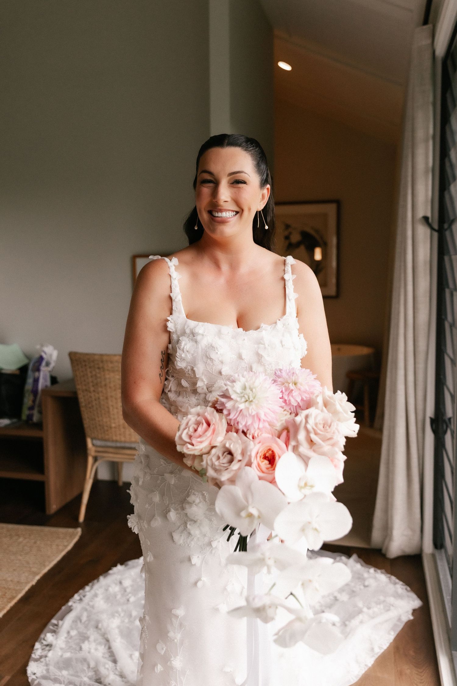 Smiling bride in white floral wedding dress holding pink and white bouquet indoors.