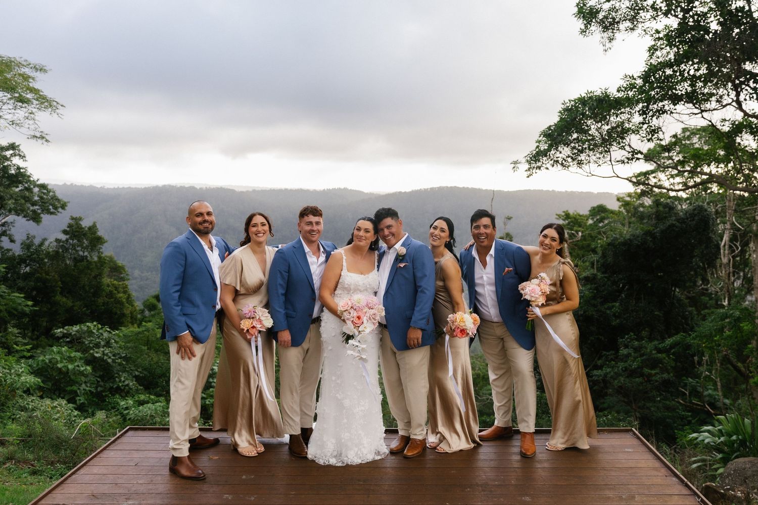 Wedding party poses on wooden deck with lush green mountain backdrop on cloudy day, bride in white gown with groomsmen and bridesmaids.