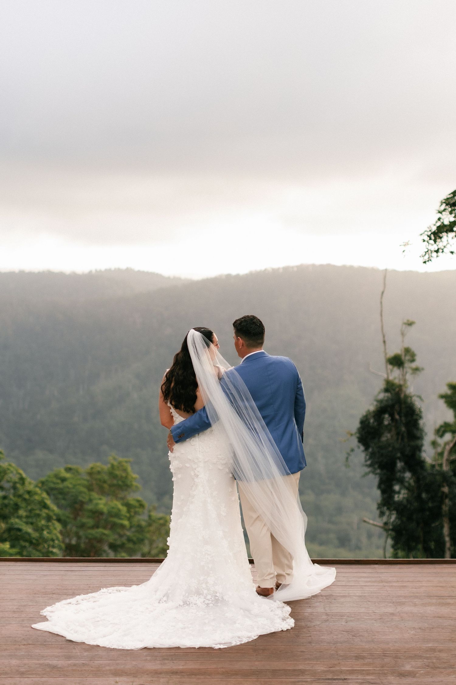 Bride and groom standing together on deck overlooking misty mountain forest landscape at sunset wedding venue.