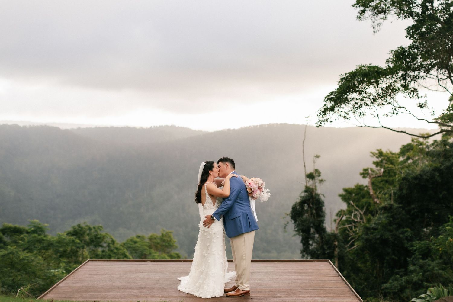 Bride and groom embrace on a wooden deck with breathtaking misty mountain rainforest views at dusk.