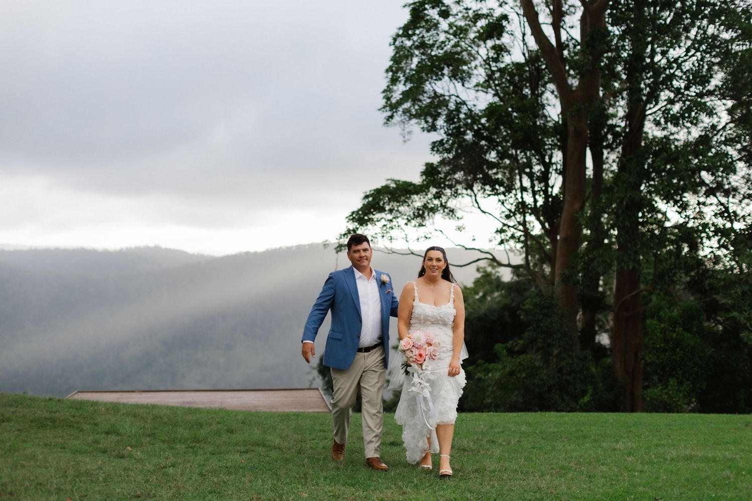 Couple walking hand in hand across green lawn with misty mountain landscape at sunset.