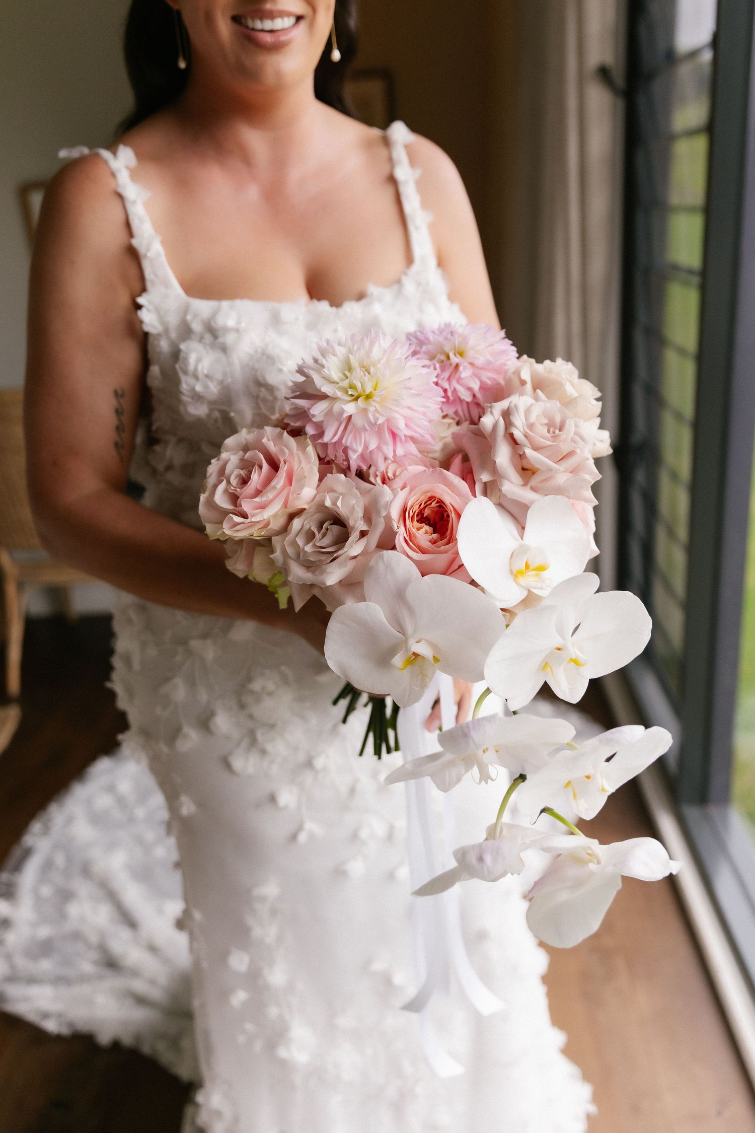 Bride in white floral gown holding elegant bouquet of pink roses, dahlias, and white orchids.