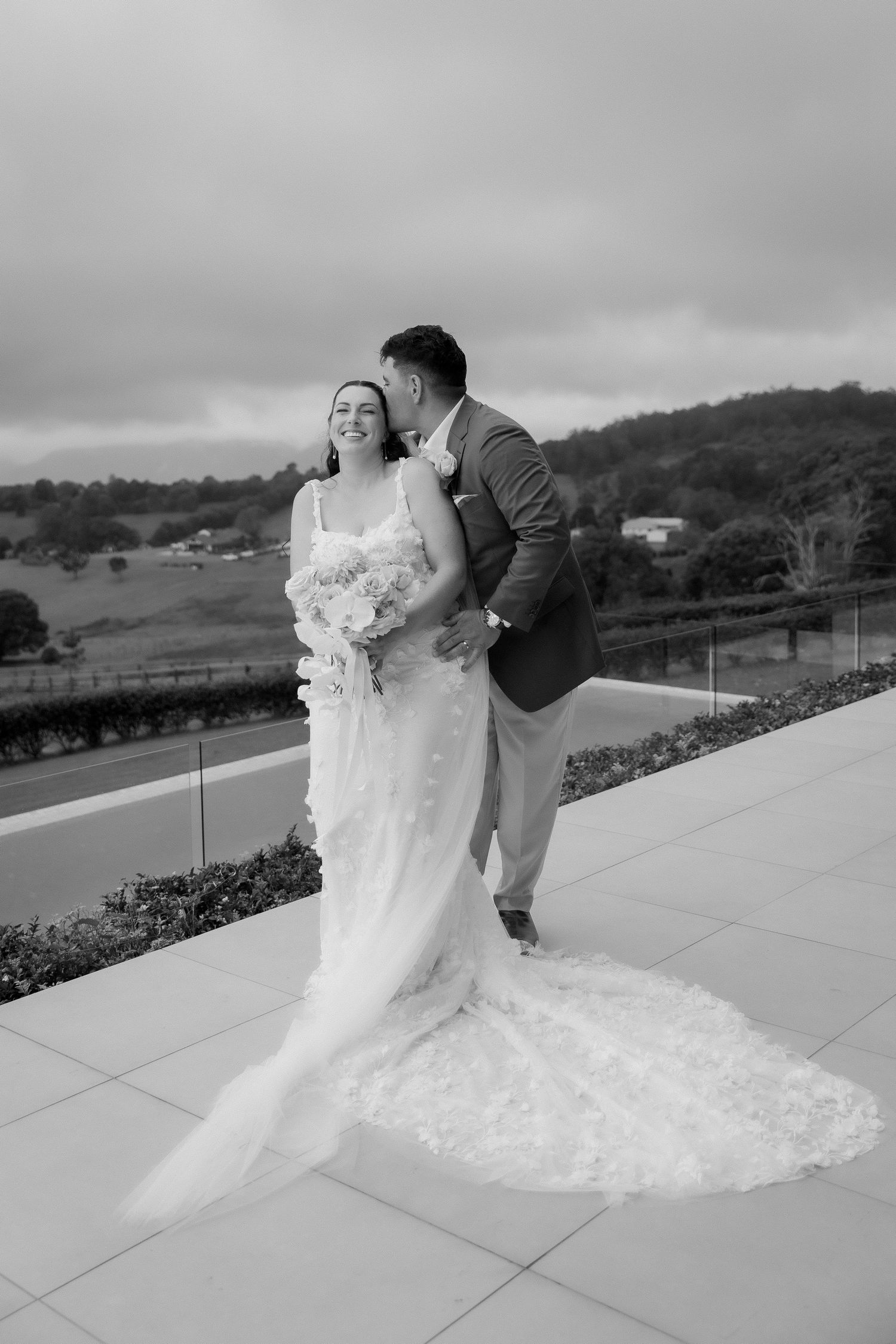 Black and white photo of groom kissing brides cheek as she smiles holding bouquet on outdoor terrace.