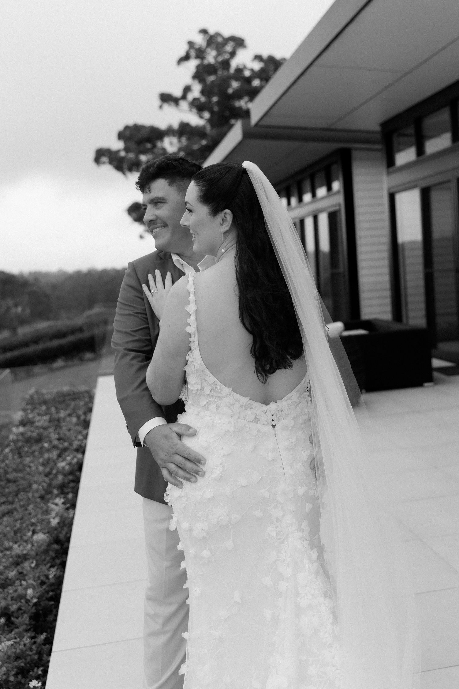 Black and white photo of bride in backless lace gown with veil and groom embracing outdoors at venue.