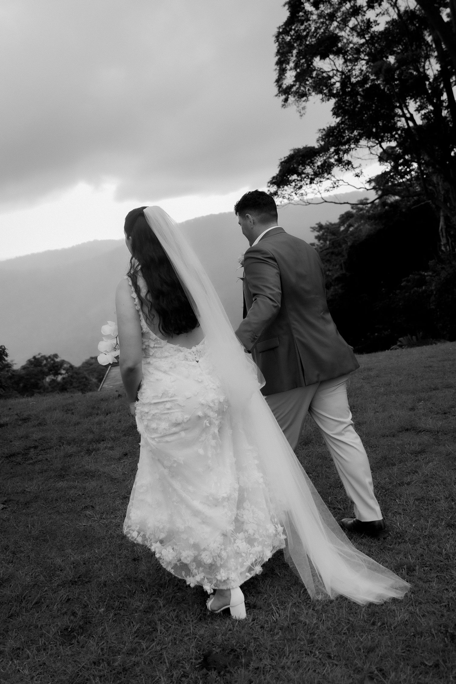 Black and white image of bride and groom walking hand in hand across grassy field with veil flowing.