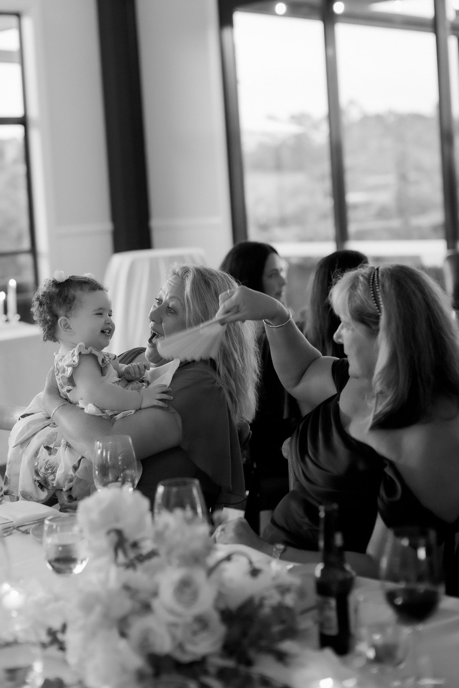 Black and white photo of guests laughing with baby at elegant indoor wedding reception.