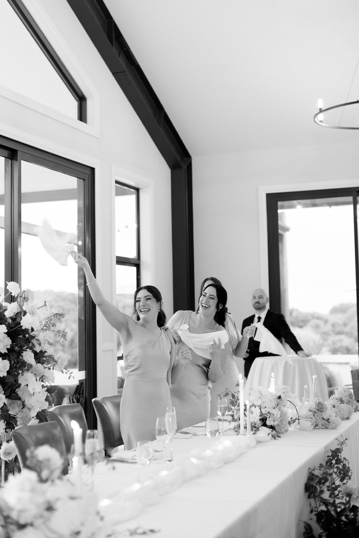 Bridesmaids celebrate at wedding reception head table, raising napkins joyfully in black and white photo.