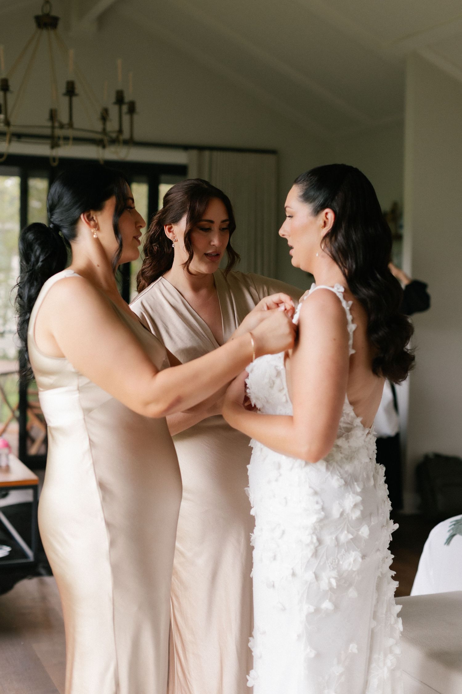 Bridesmaids in champagne gowns helping bride into her white floral wedding dress.