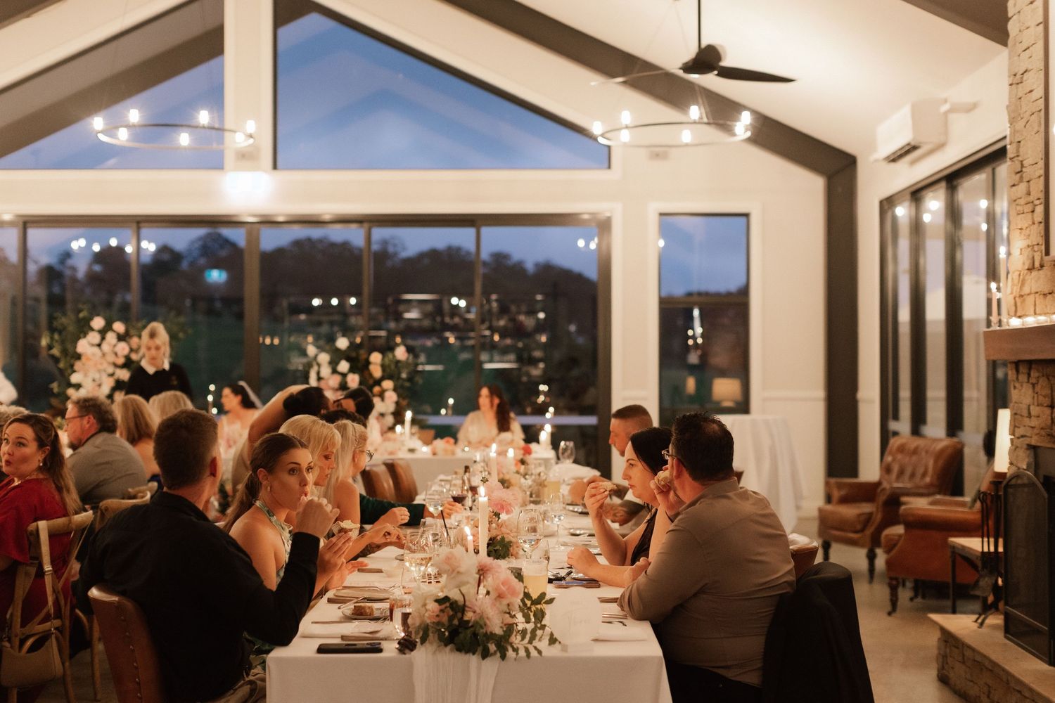 Elegant wedding reception dinner in modern venue with large windows, candles, and floral centerpieces at dusk.