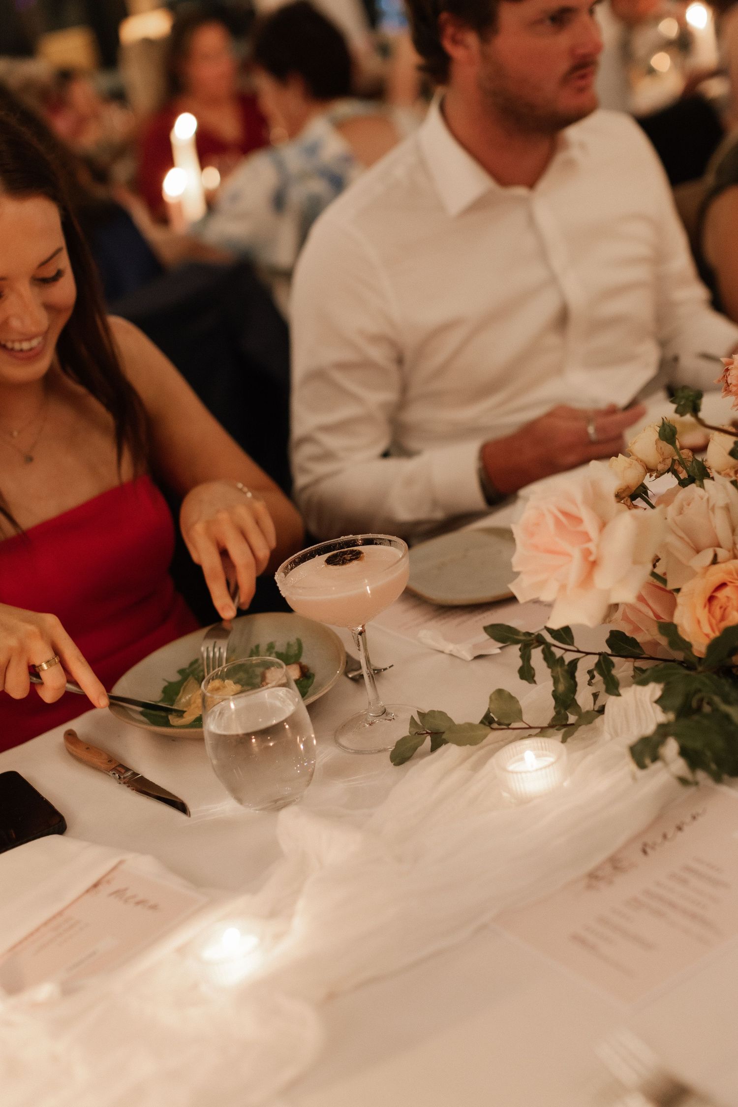 Wedding reception guest reaching for a cocktail beside a floral centerpiece with soft candlelight.