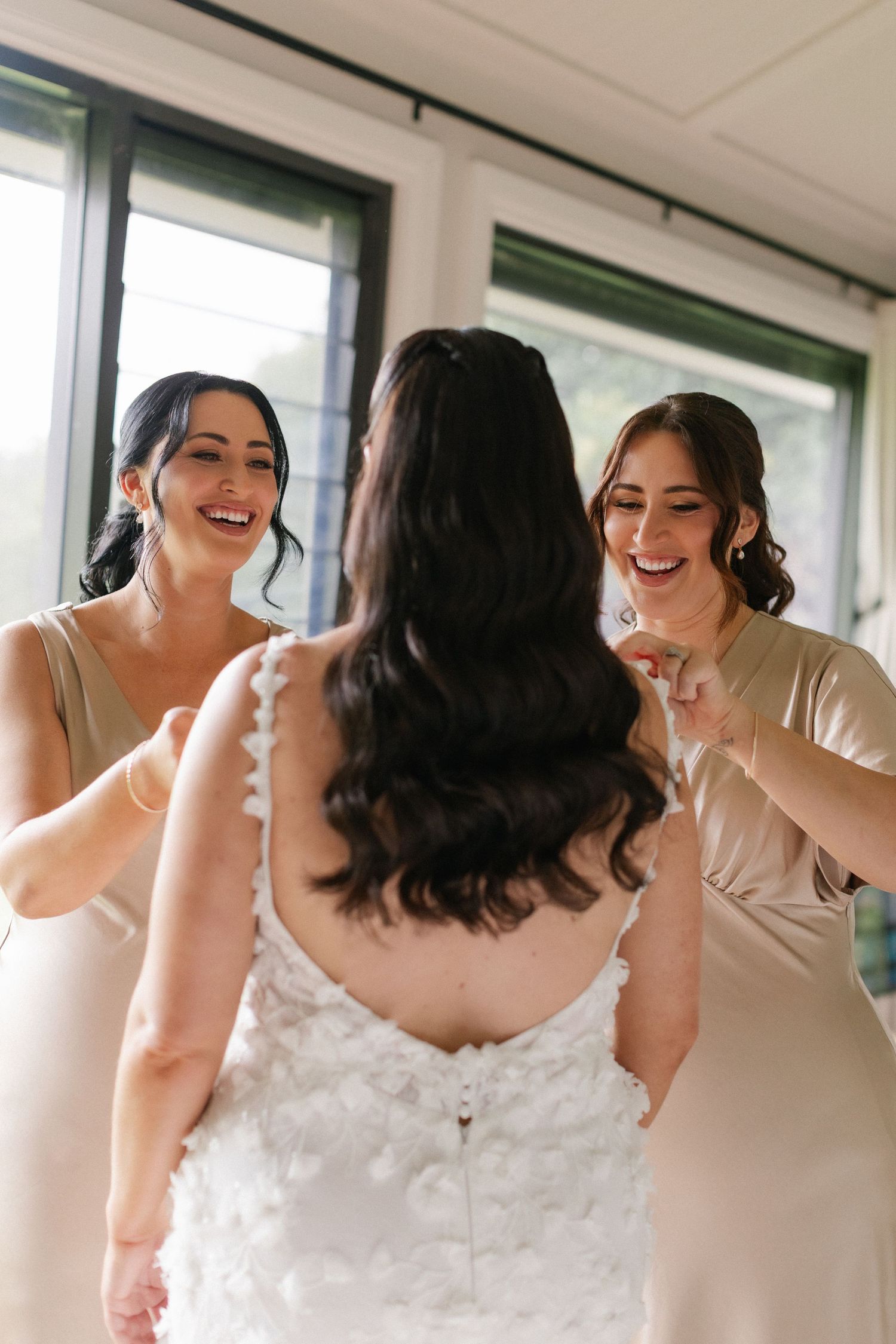 Two smiling bridesmaids in champagne dresses help bride with her white floral wedding gown indoors.