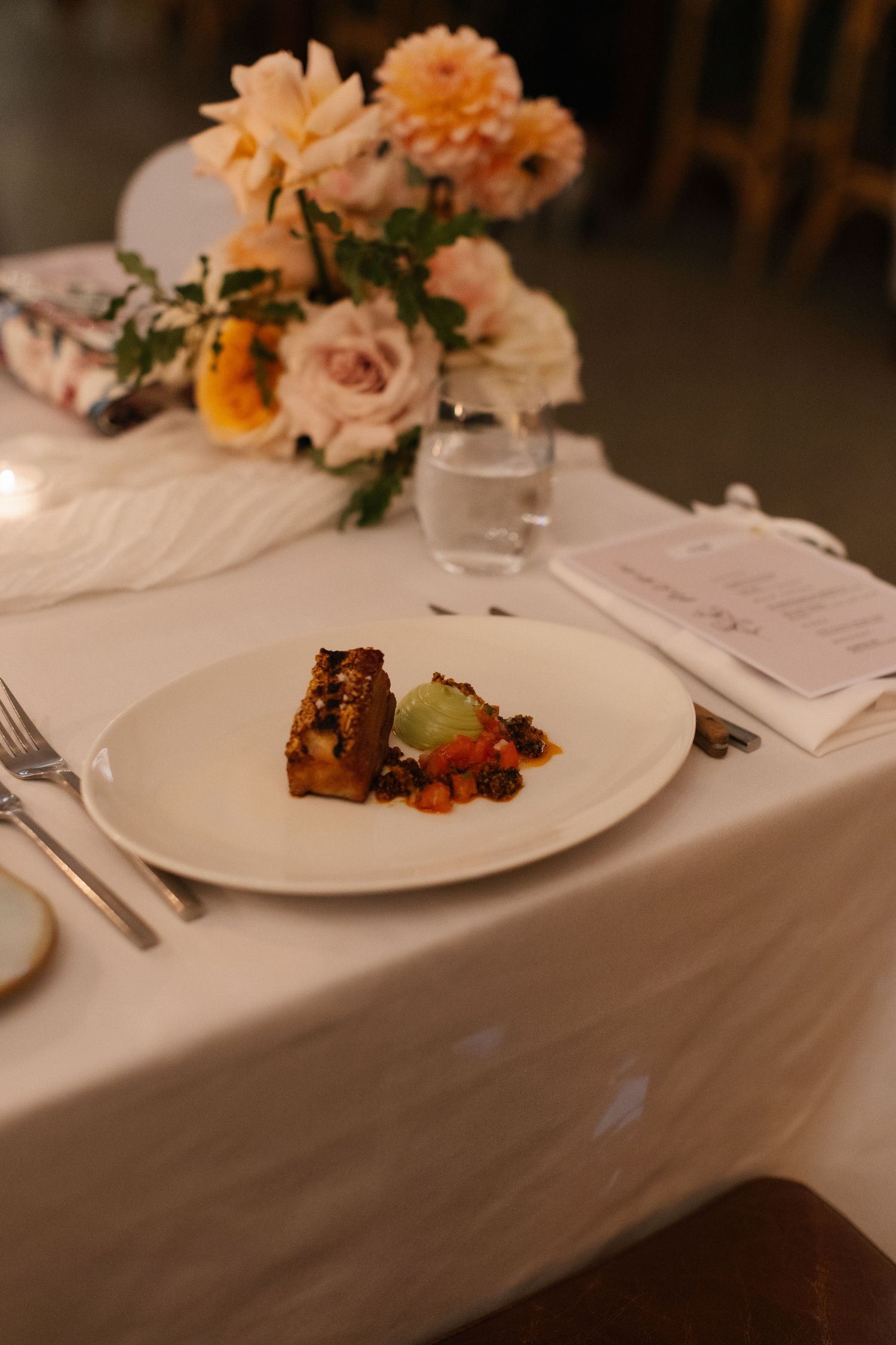 Elegant wedding dinner plate with gourmet dish served on white tablecloth beside peach floral centerpiece and candle.