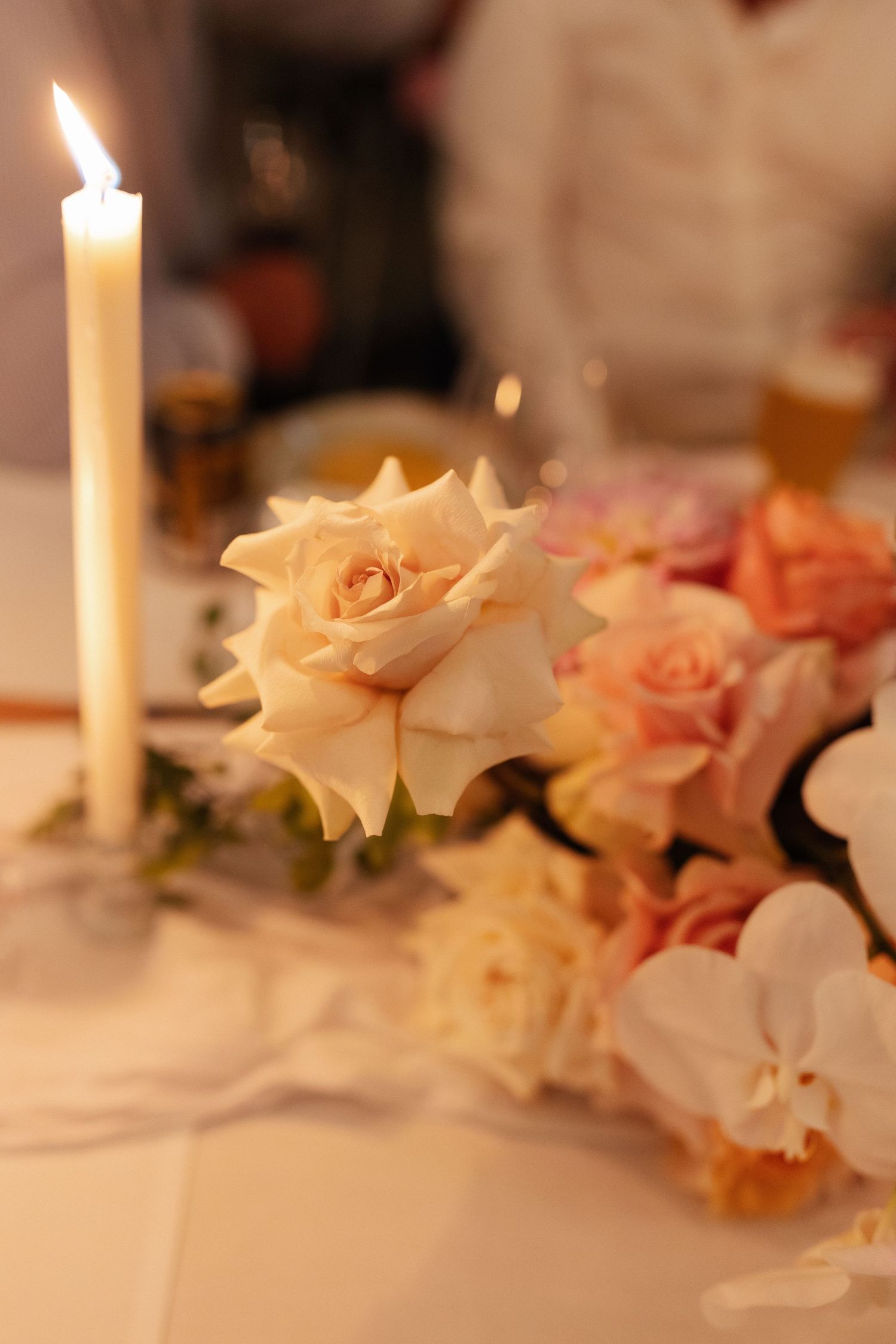 Lit white taper candle beside cream and pink roses centerpiece at wedding reception.
