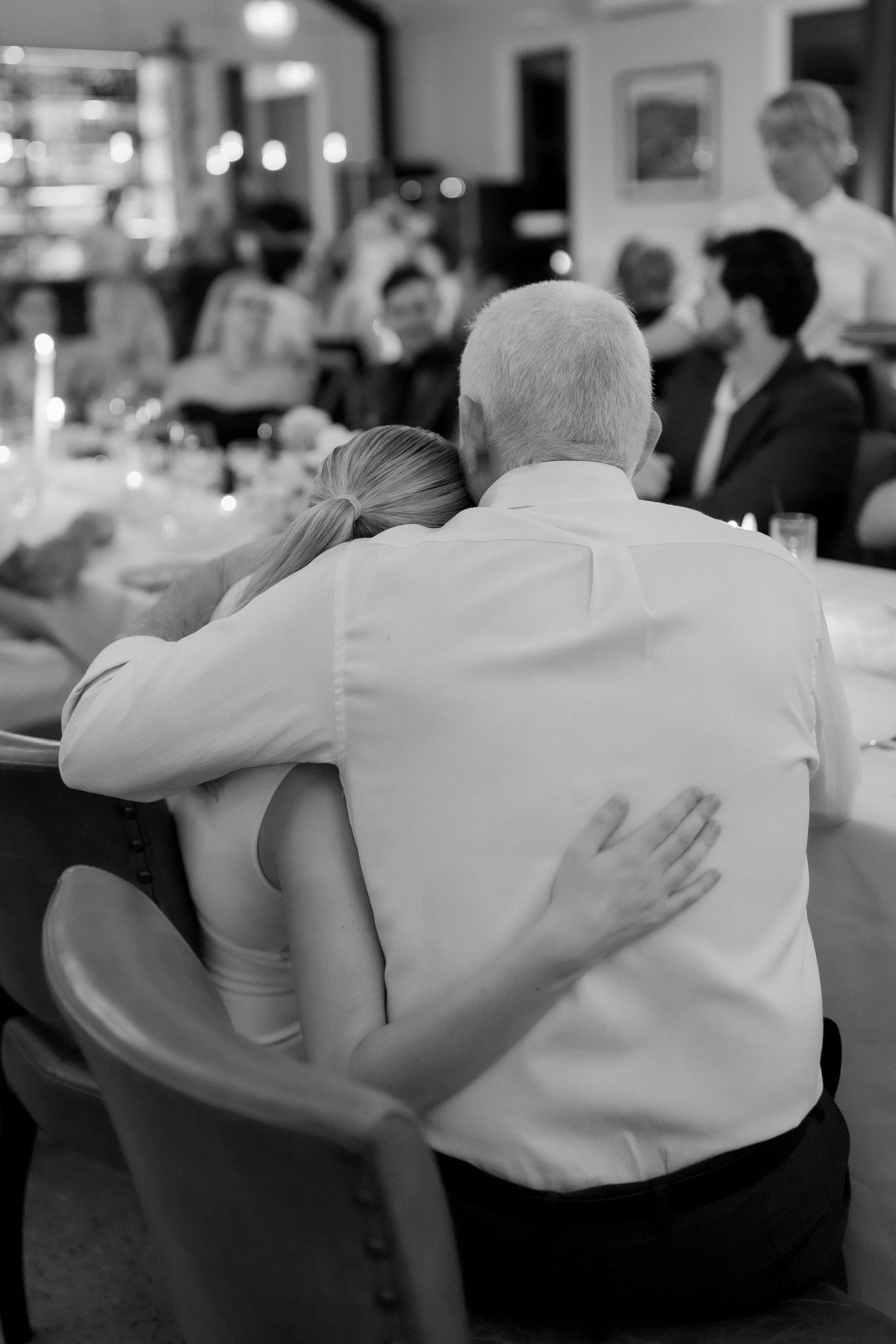 Black and white image of woman resting head on older mans shoulder at wedding reception.