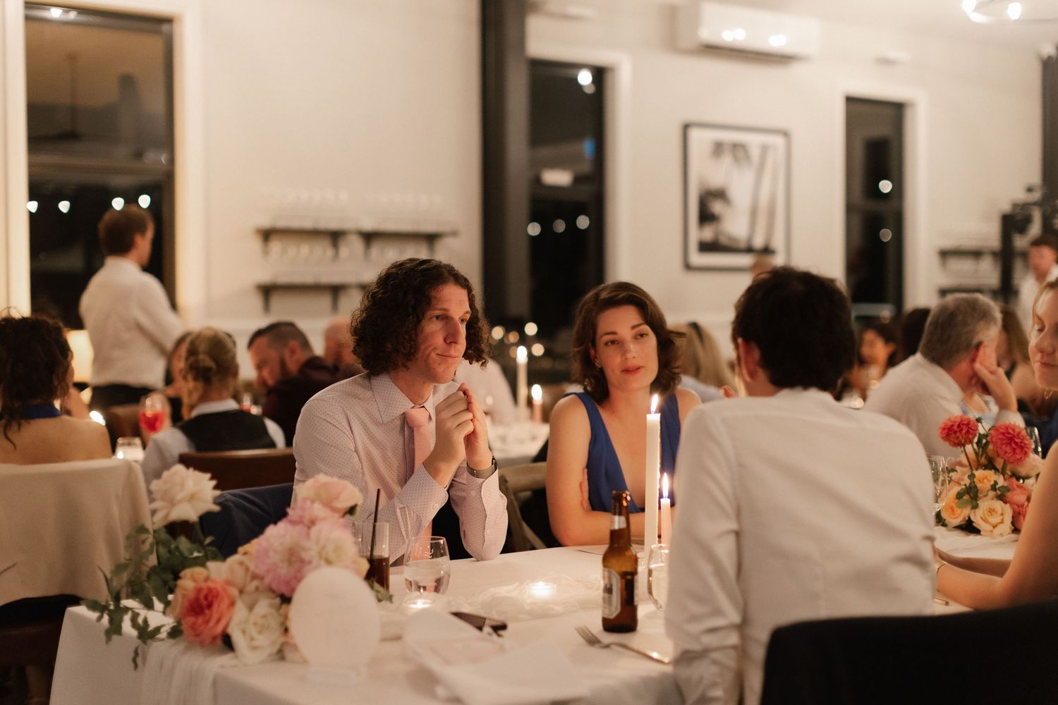 Wedding guests enjoying candlelit dinner reception with floral centerpieces in elegant venue.