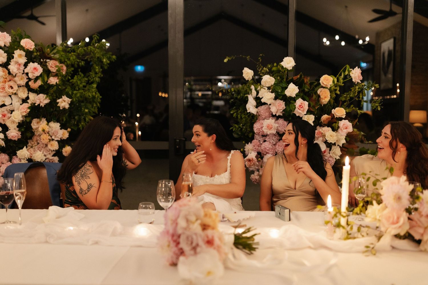 Bridal party seated at elegant reception table with pink floral arrangements and candlelight indoors.