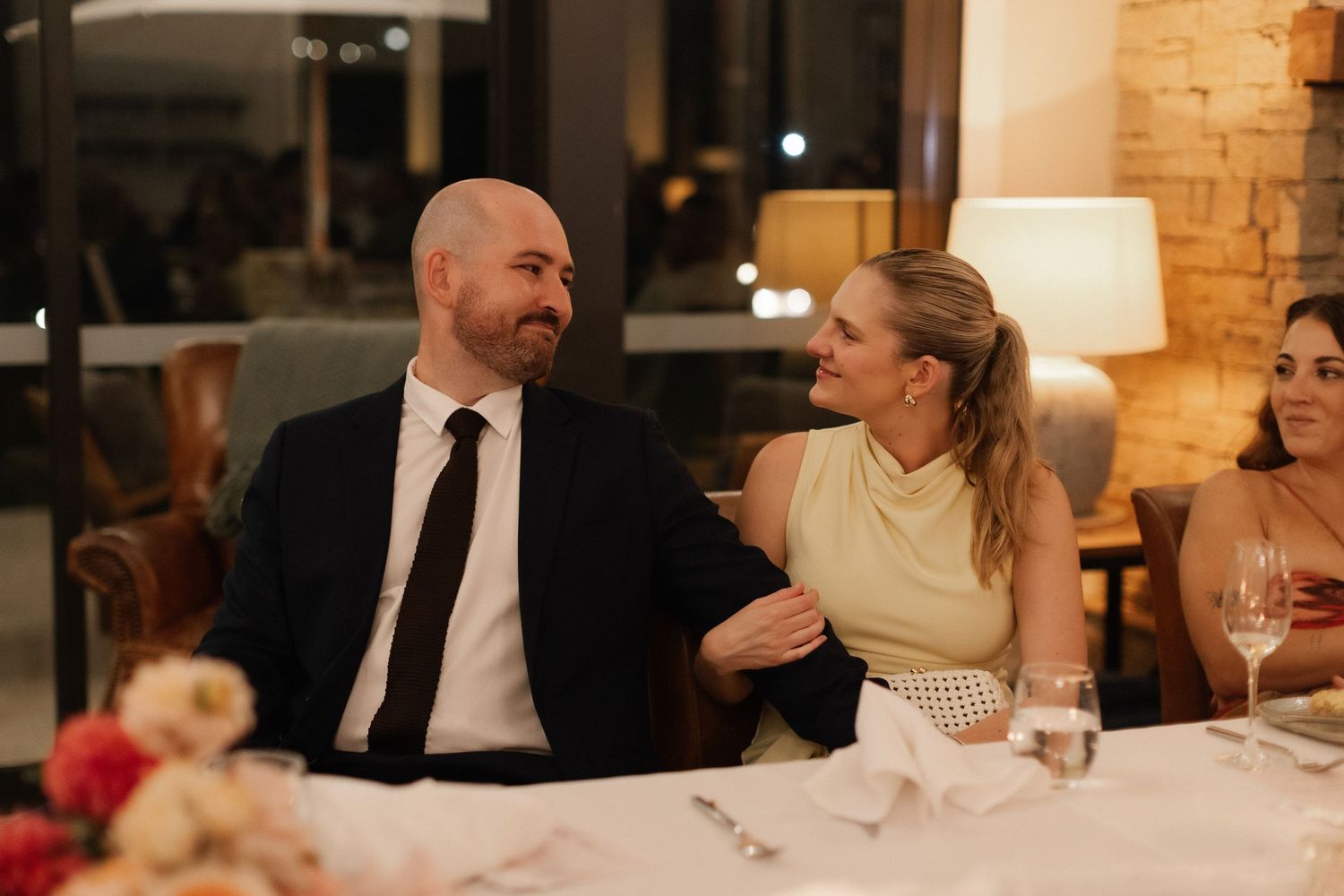 Couple in formal attire smiling at each other while seated at elegant wedding reception dinner table.