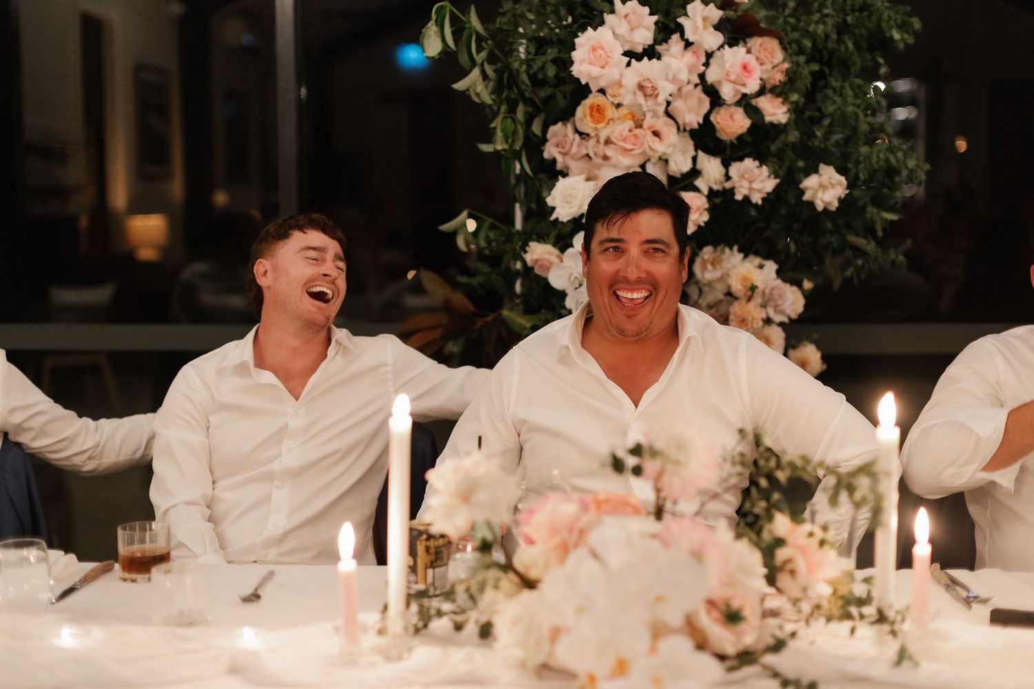 Two men in white shirts laughing joyfully at a candlelit wedding reception table with pink floral arrangements.