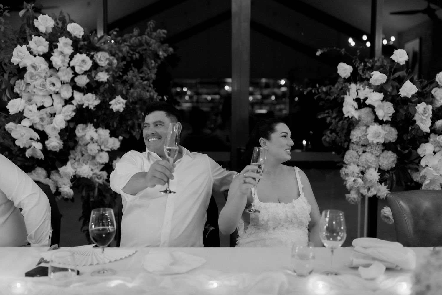 Bride and groom toasting champagne at head table with white floral arrangements.