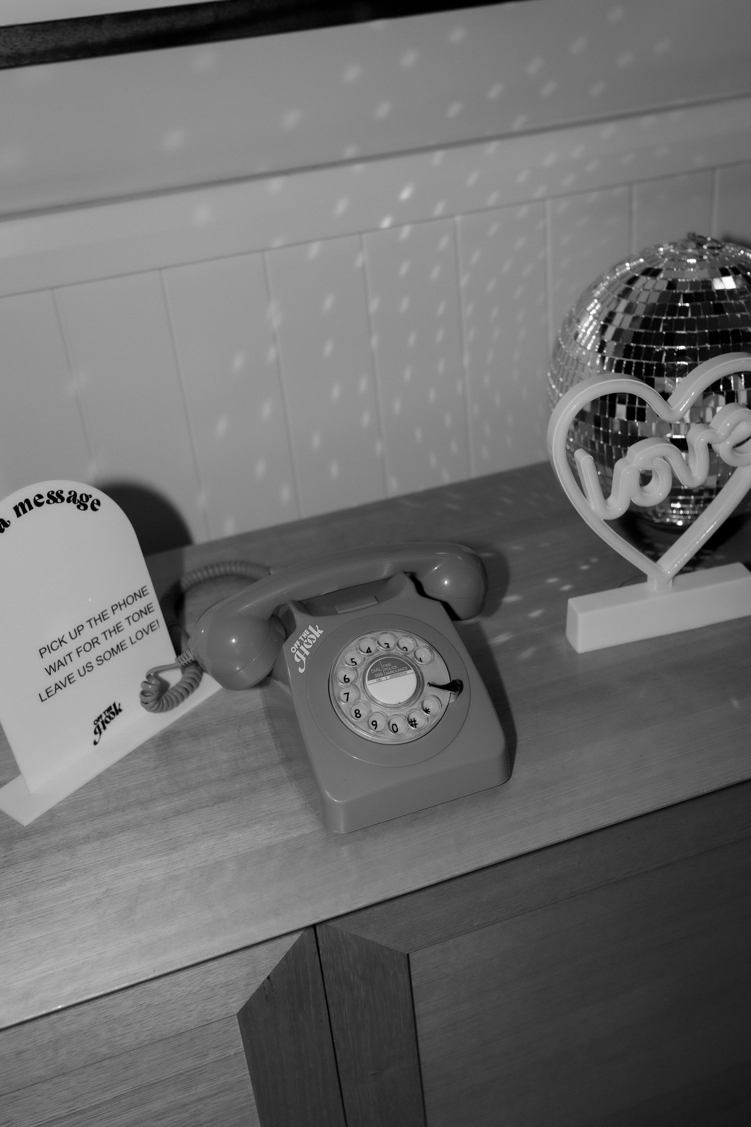 Black and white photo of a vintage rotary phone, love heart sign, and disco ball on a wooden shelf.