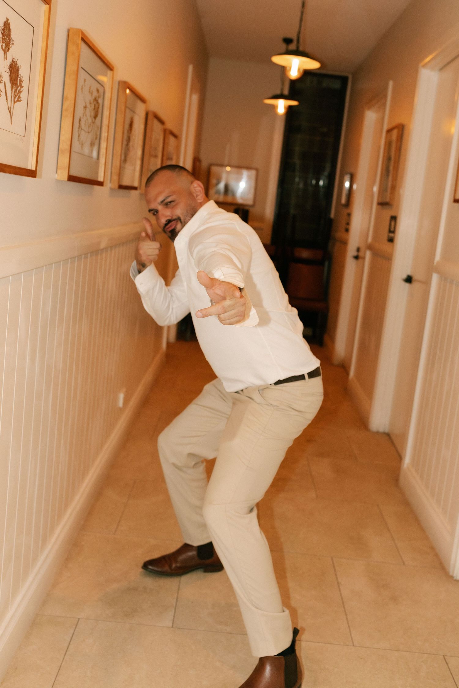 Man in white shirt and khaki pants posing playfully in a hallway with framed artwork on the walls.