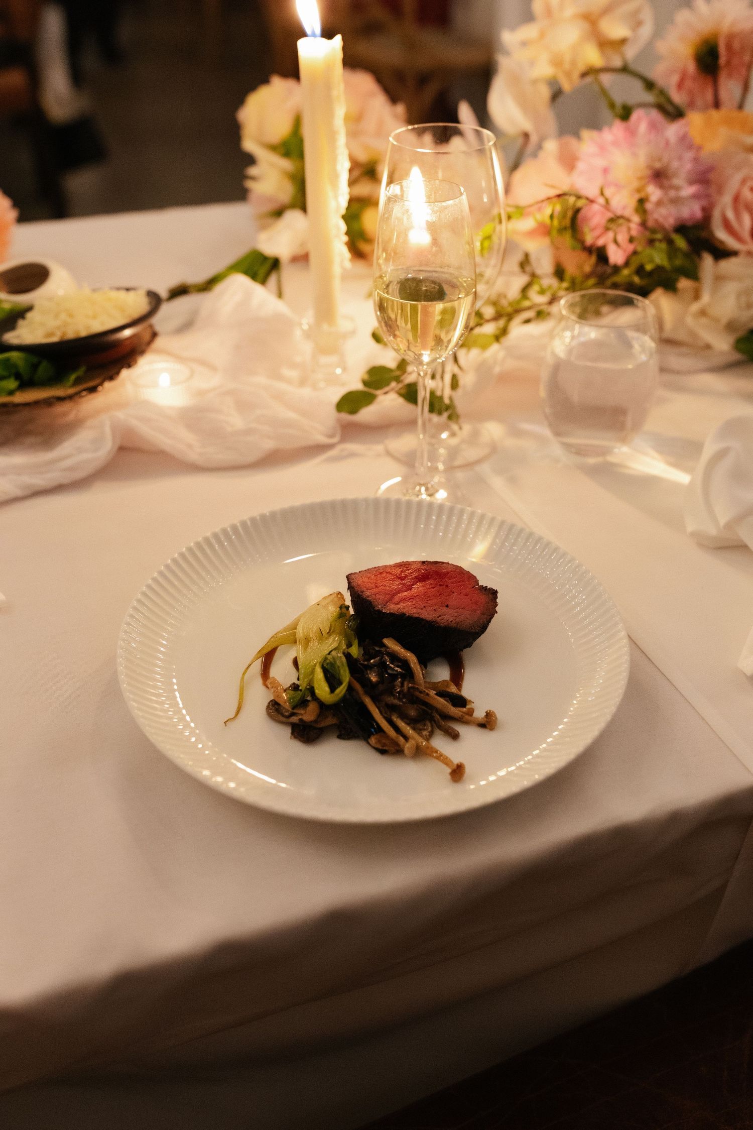 Elegant wedding reception dinner plate with seared beef and mushrooms beside candlelight and floral centerpiece.