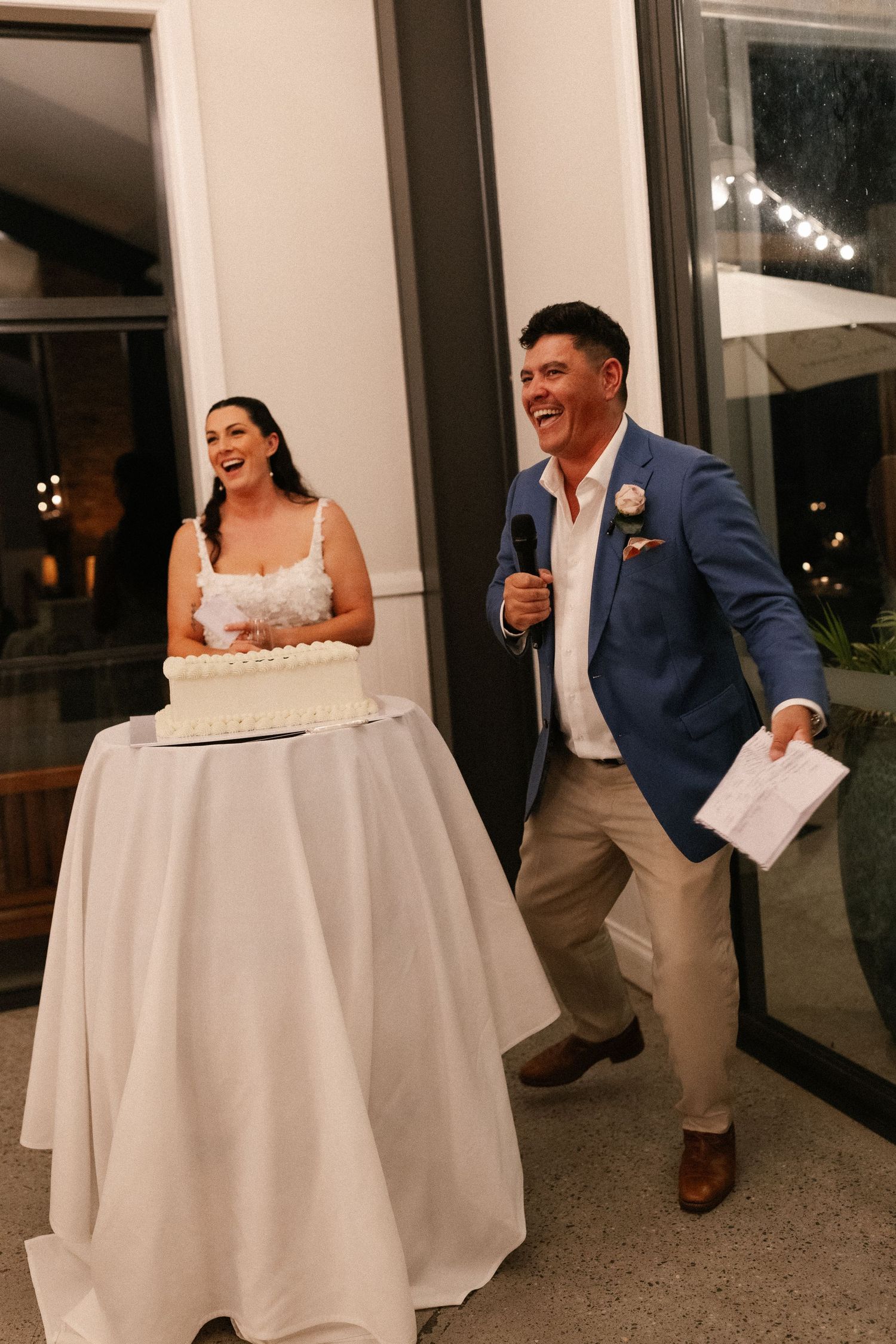 Laughing bride and groom in blue blazer giving speech beside white wedding cake at evening reception.