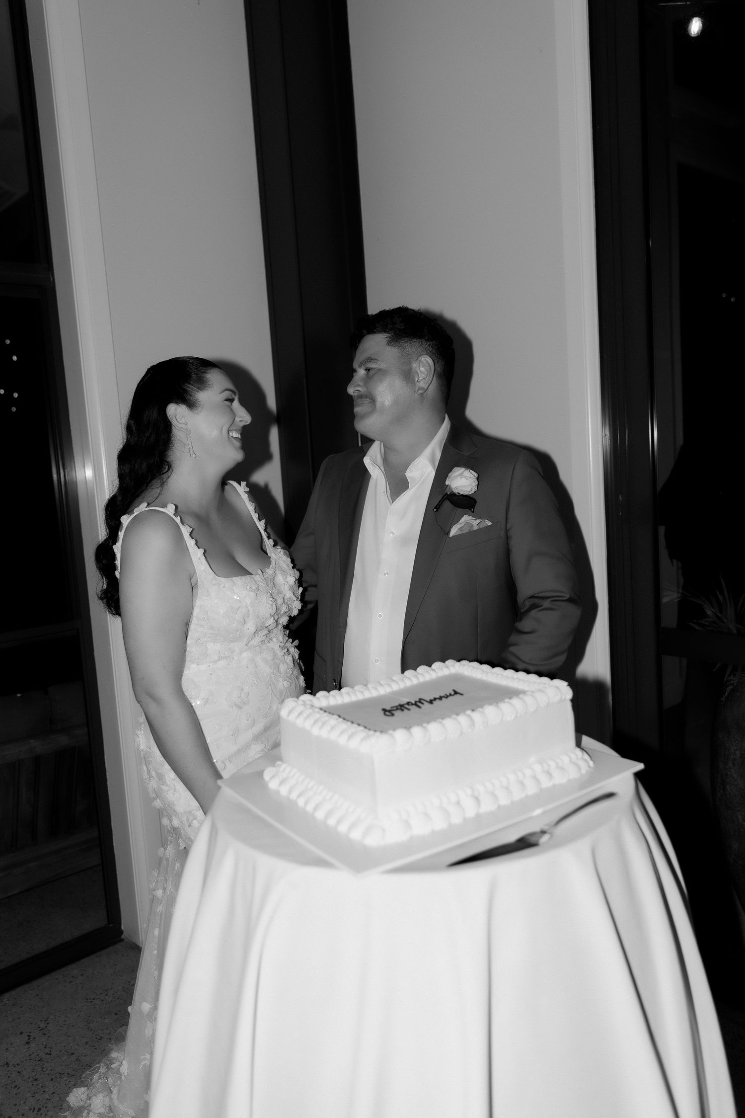 Black and white photo of smiling bride and groom standing beside their decorated wedding cake.