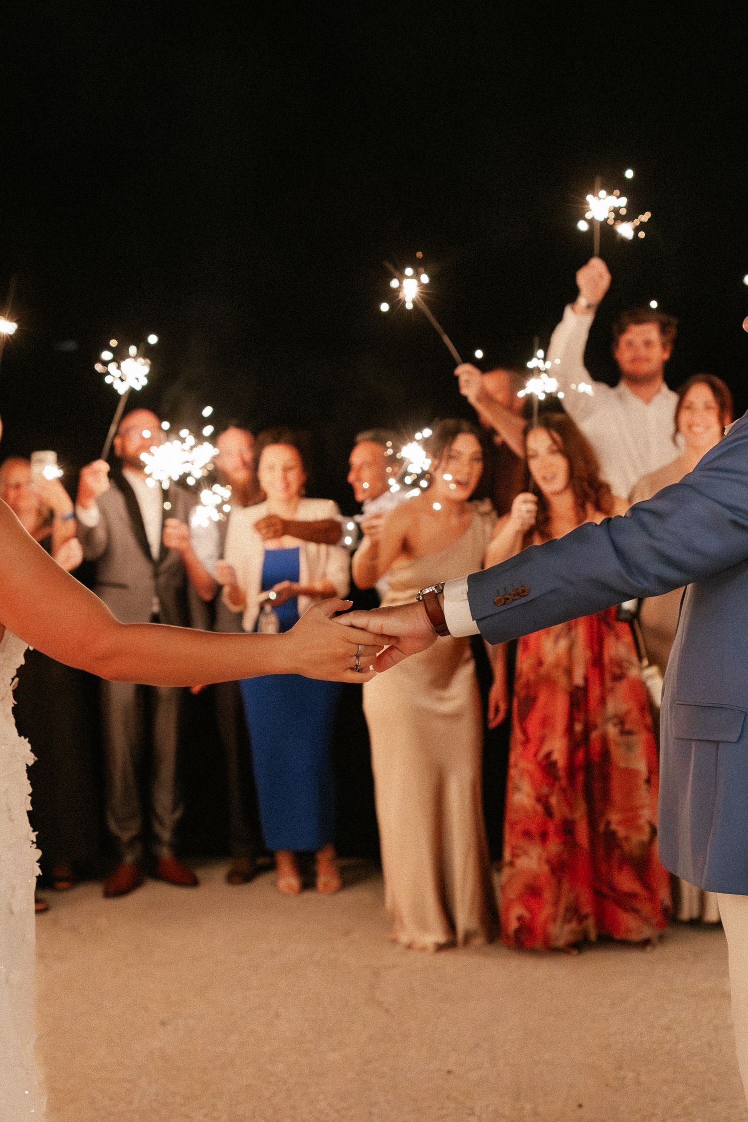 Bride and groom hold hands during a romantic nighttime wedding sparkler send-off with guests celebrating around them.