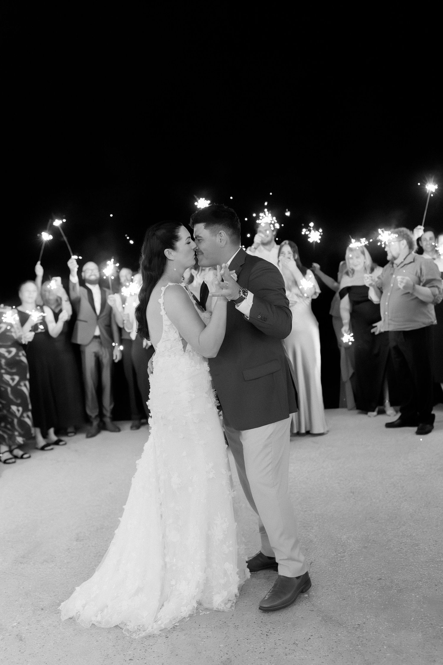 Black and white photo of bride and groom kissing surrounded by wedding guests holding sparklers at night.