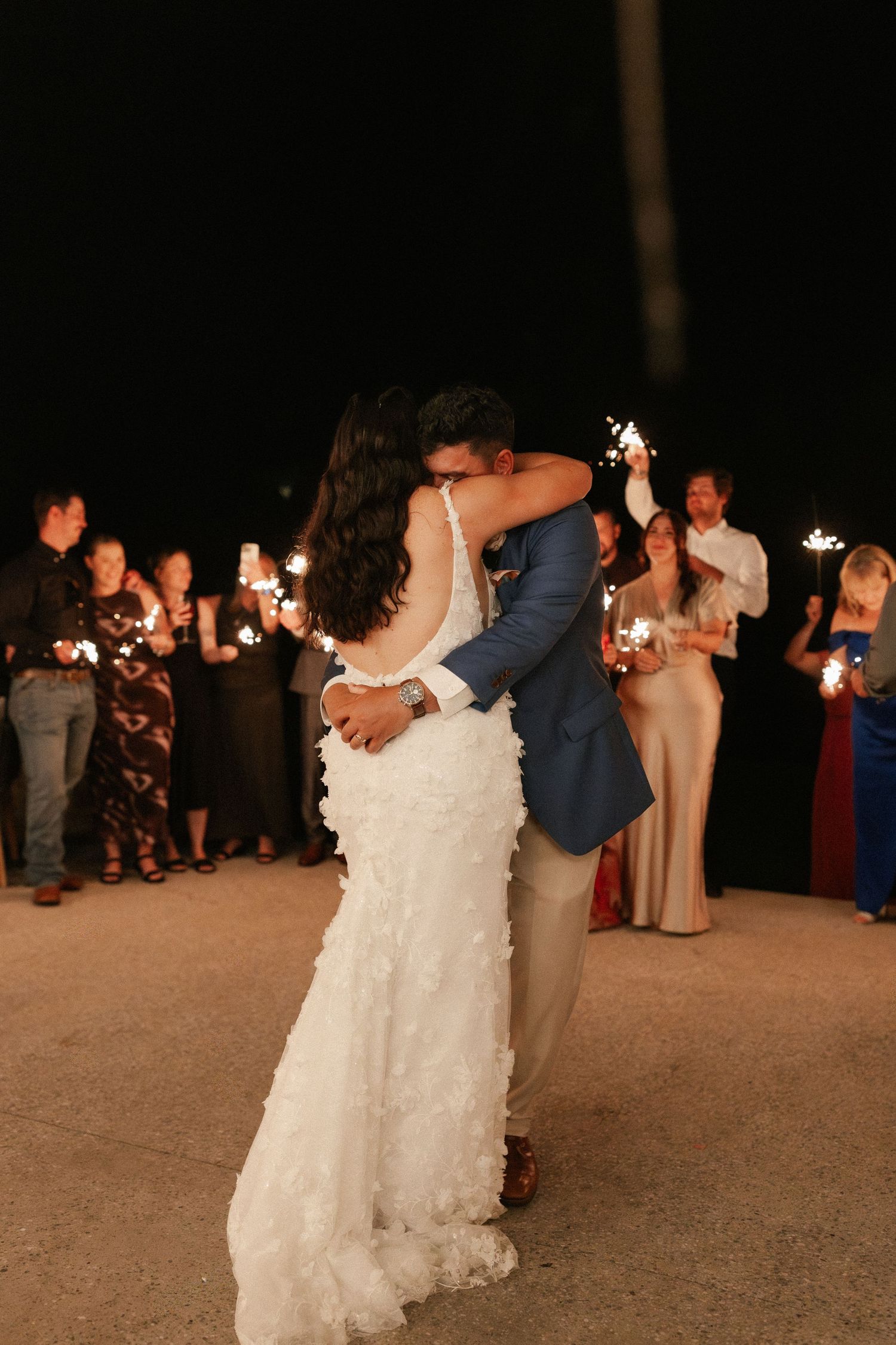 Bride and groom share romantic kiss while wedding guests hold sparklers in the background at night.