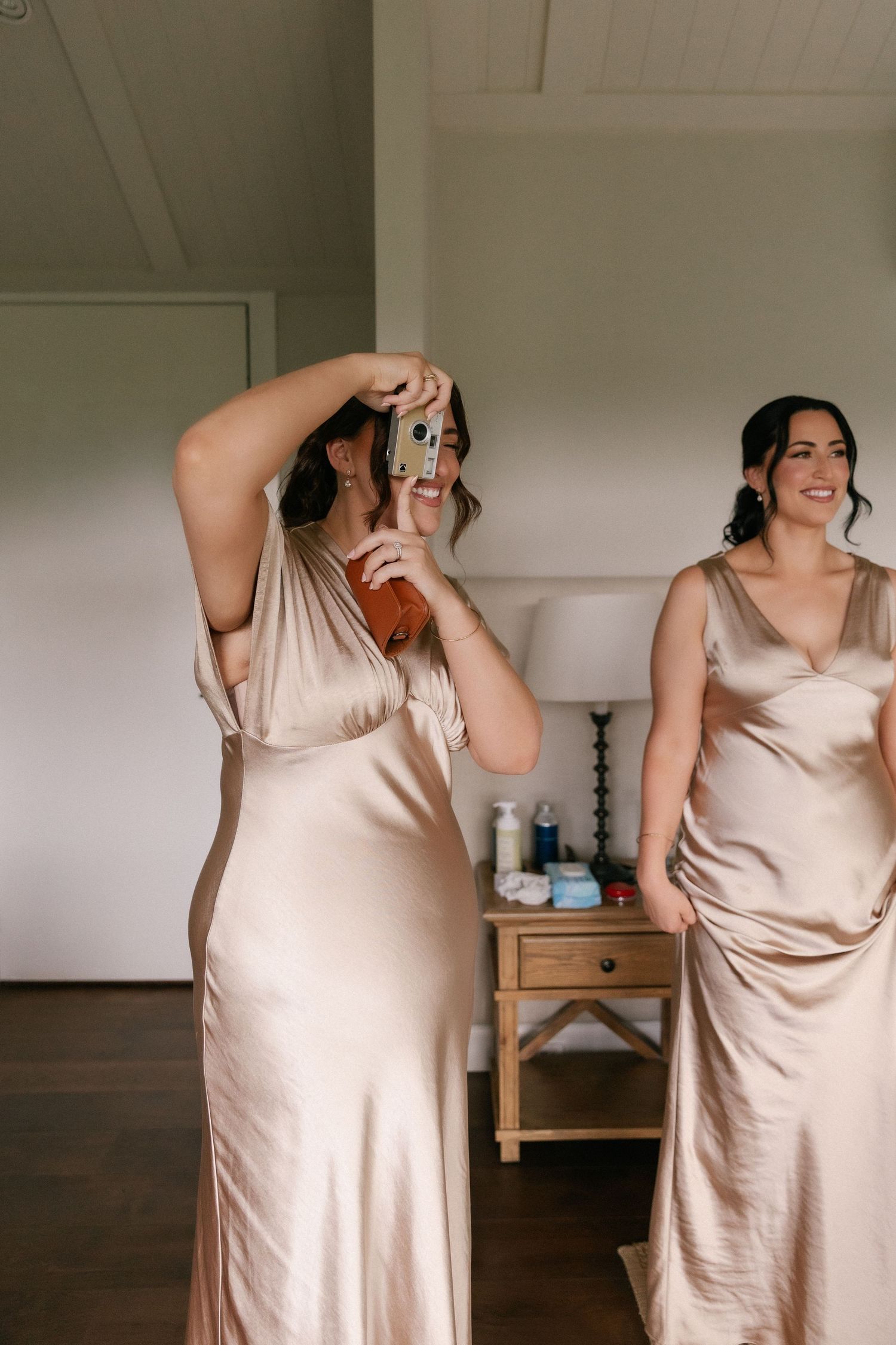 Two bridesmaids in champagne satin gowns, one taking a photo with a camera indoors.