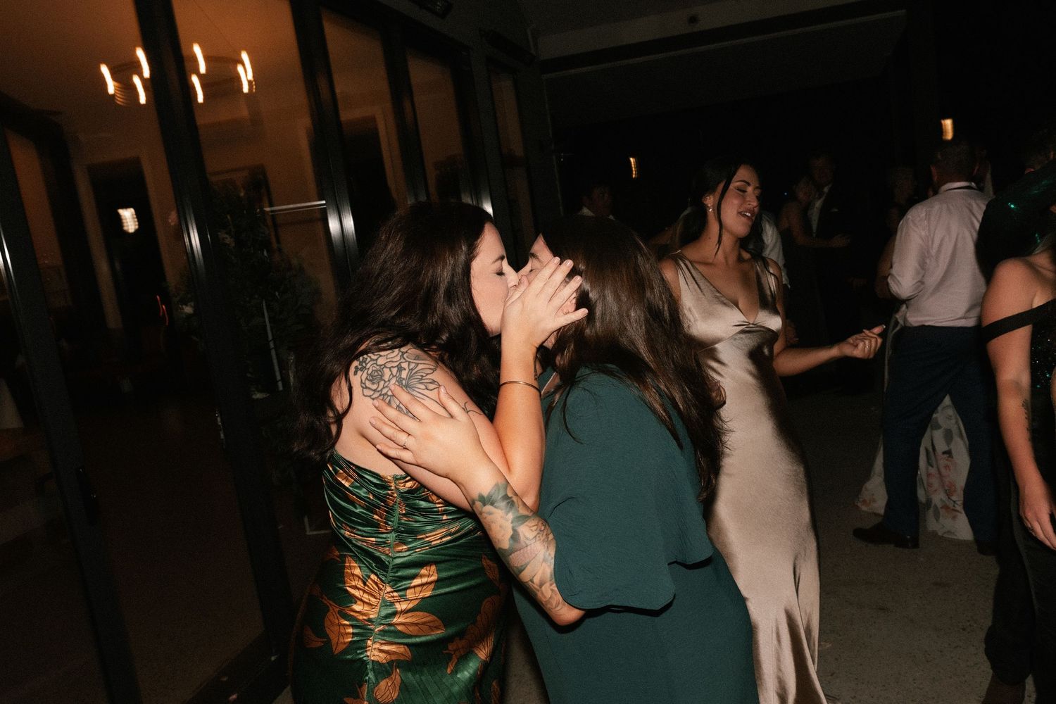 Two women share a kiss on a dimly lit wedding reception dance floor.