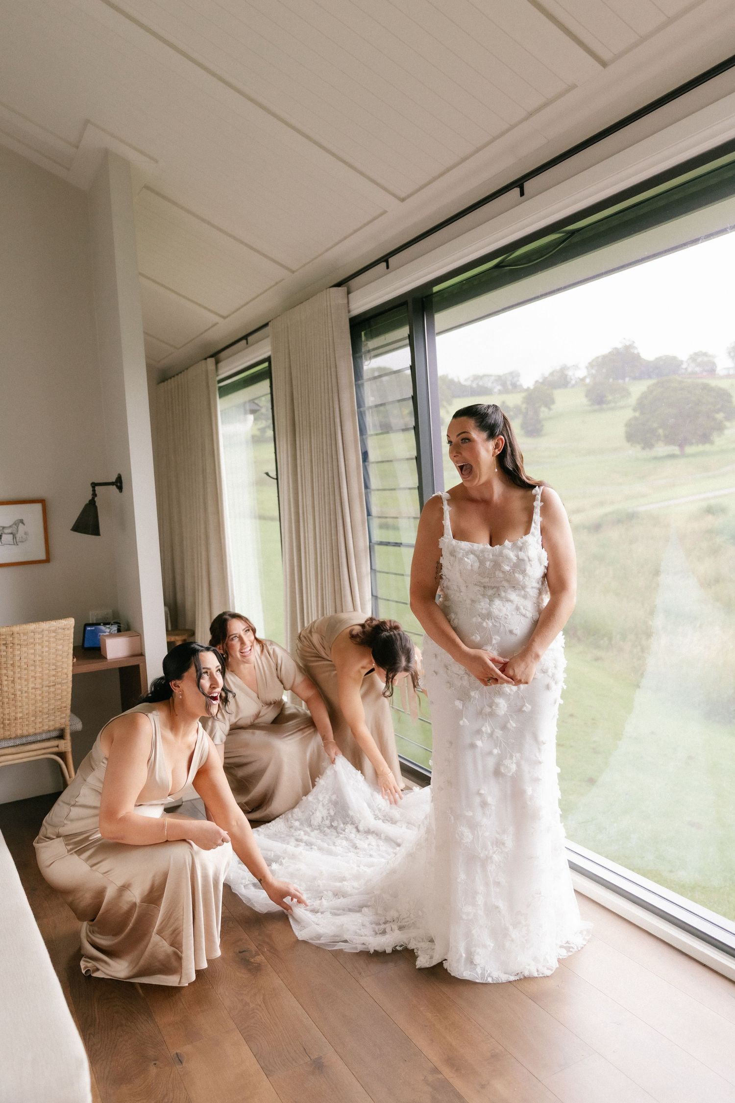 Bride in floral white gown stands by large windows as bridesmaids in champagne dresses adjust her train.