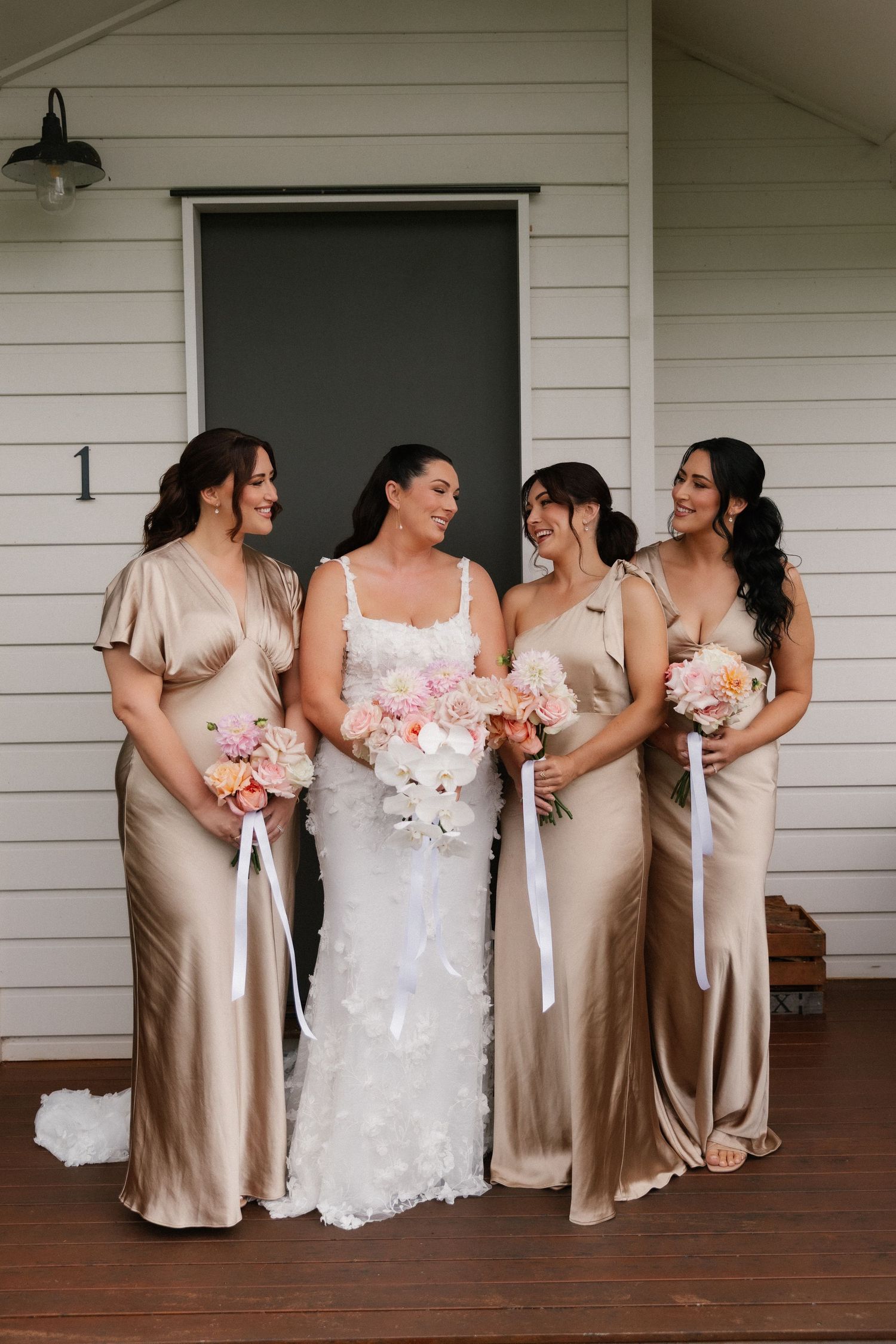 Bride in lace gown with three bridesmaids in champagne satin dresses holding pink and white bouquets.