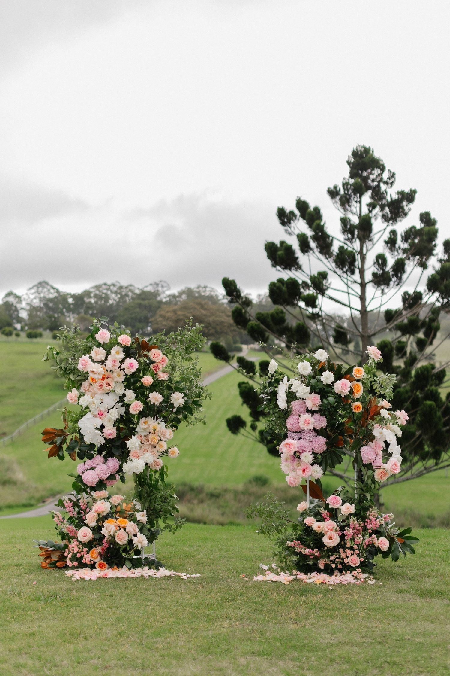 Two floral arches adorned with pink and white blooms stand on a green lawn with rolling hills and trees in the background.